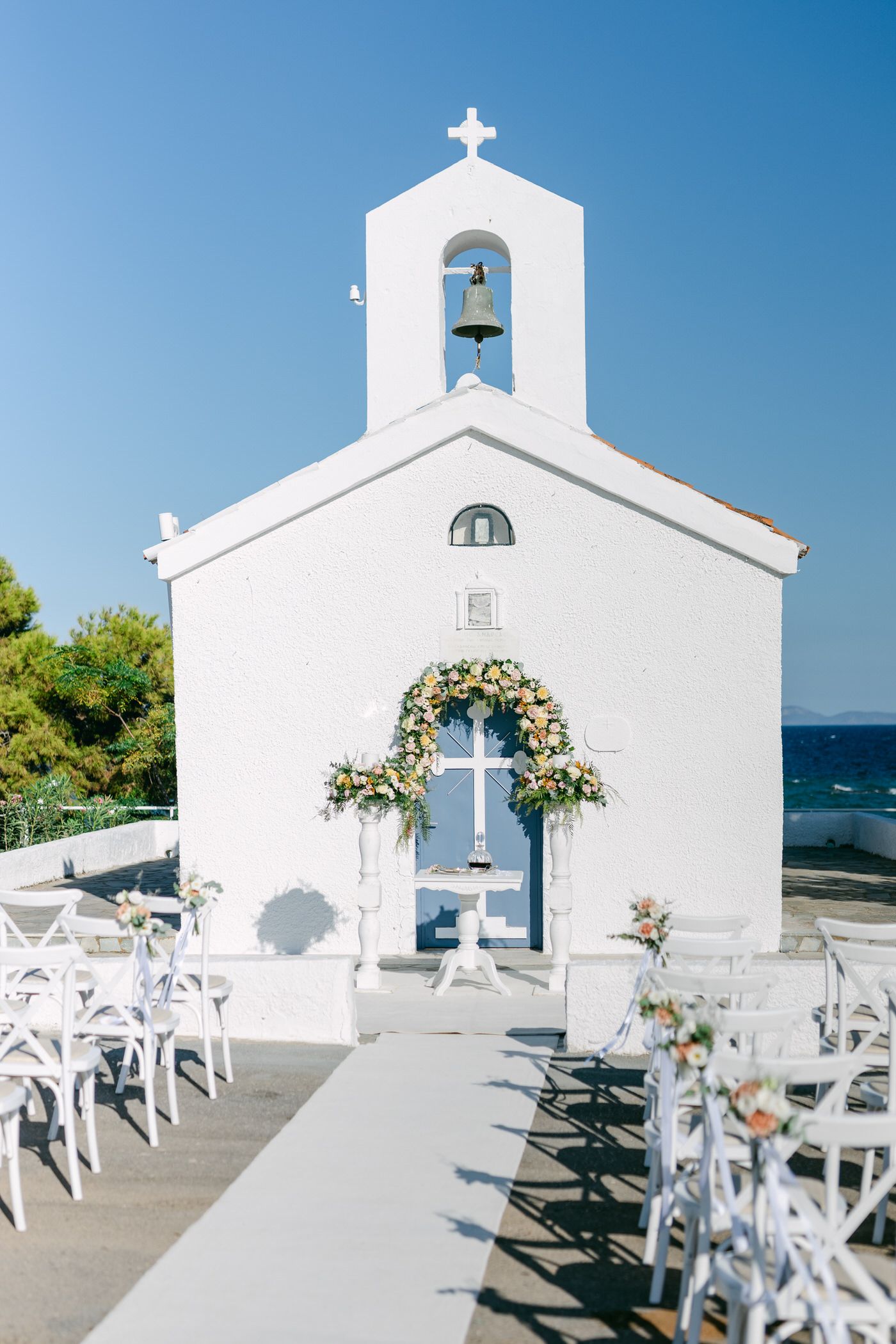 Small white Greek chapel with floral arch and cross set up for seaside wedding ceremony with white chairs in Greece.