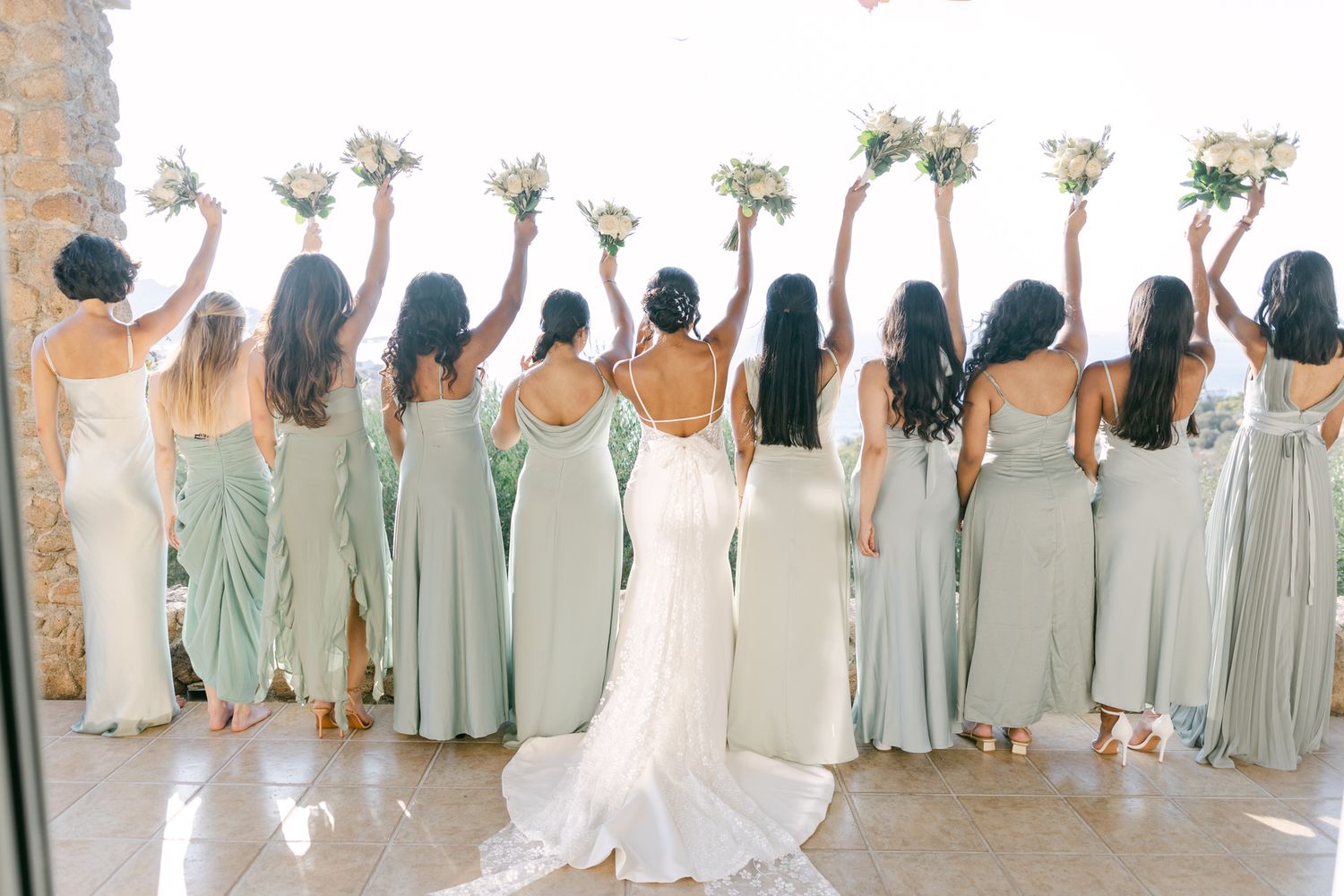 Bridesmaids in sage green dresses stand with their backs turned holding bouquets next to bride in white gown.