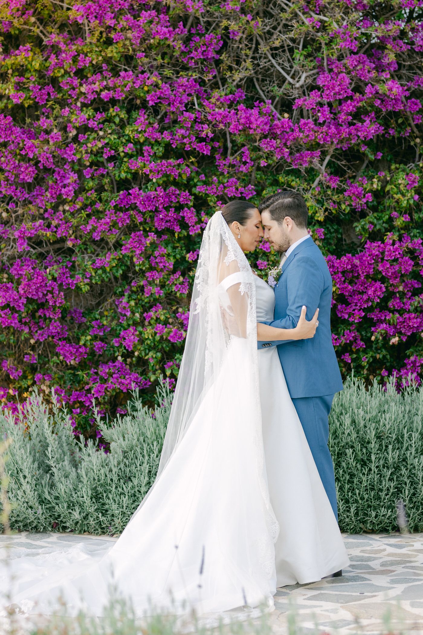 A wedding couple shares a romantic moment against a backdrop of vibrant purple bougainvillea flowers and greenery.