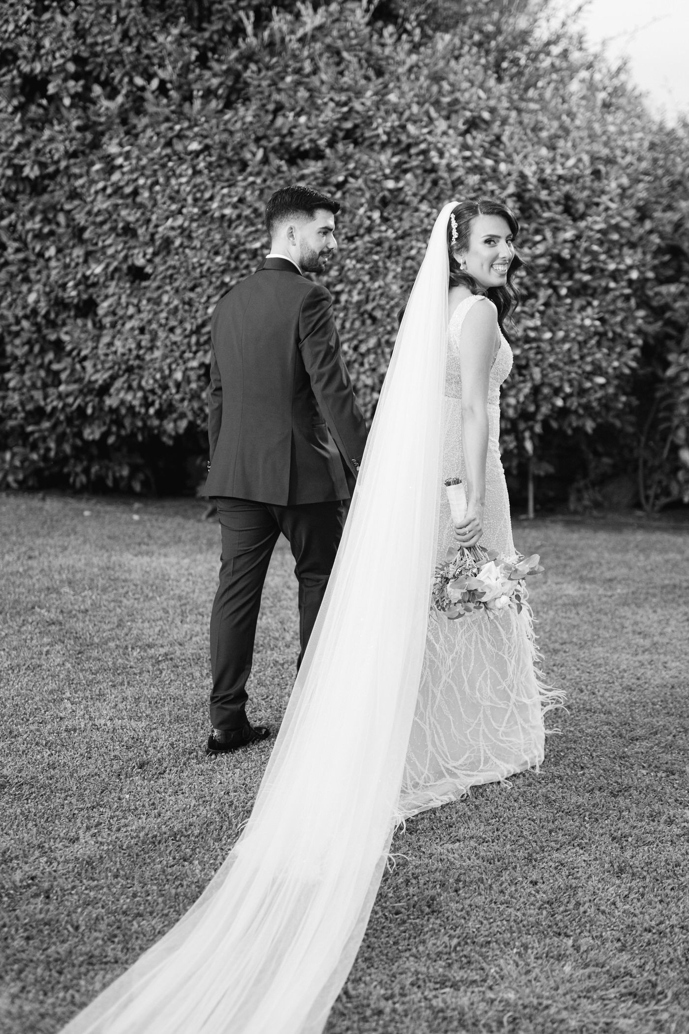 A black and white wedding photo showing a couple standing back to back outdoors with the bride wearing a long flowing veil.