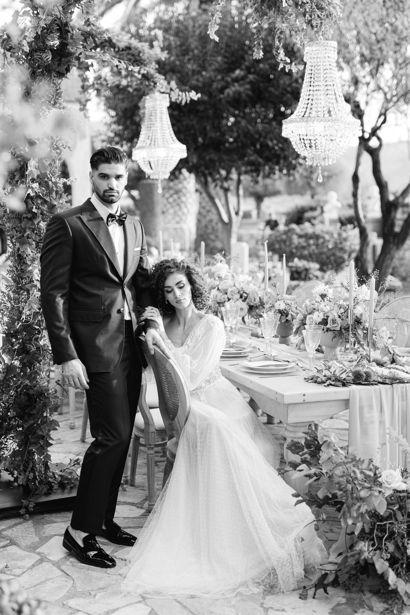 Black-and-white portrait of a bride seated at a styled reception table with the groom standing beside her under chandeliers.