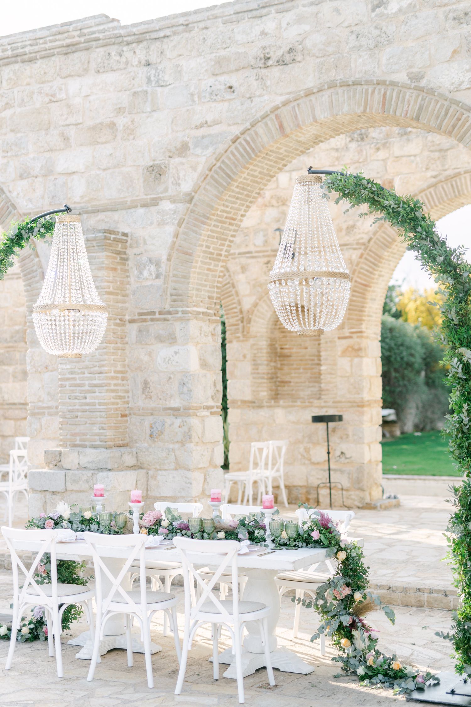 Elegant outdoor wedding reception with crystal chandeliers and white chairs under stone arches.
