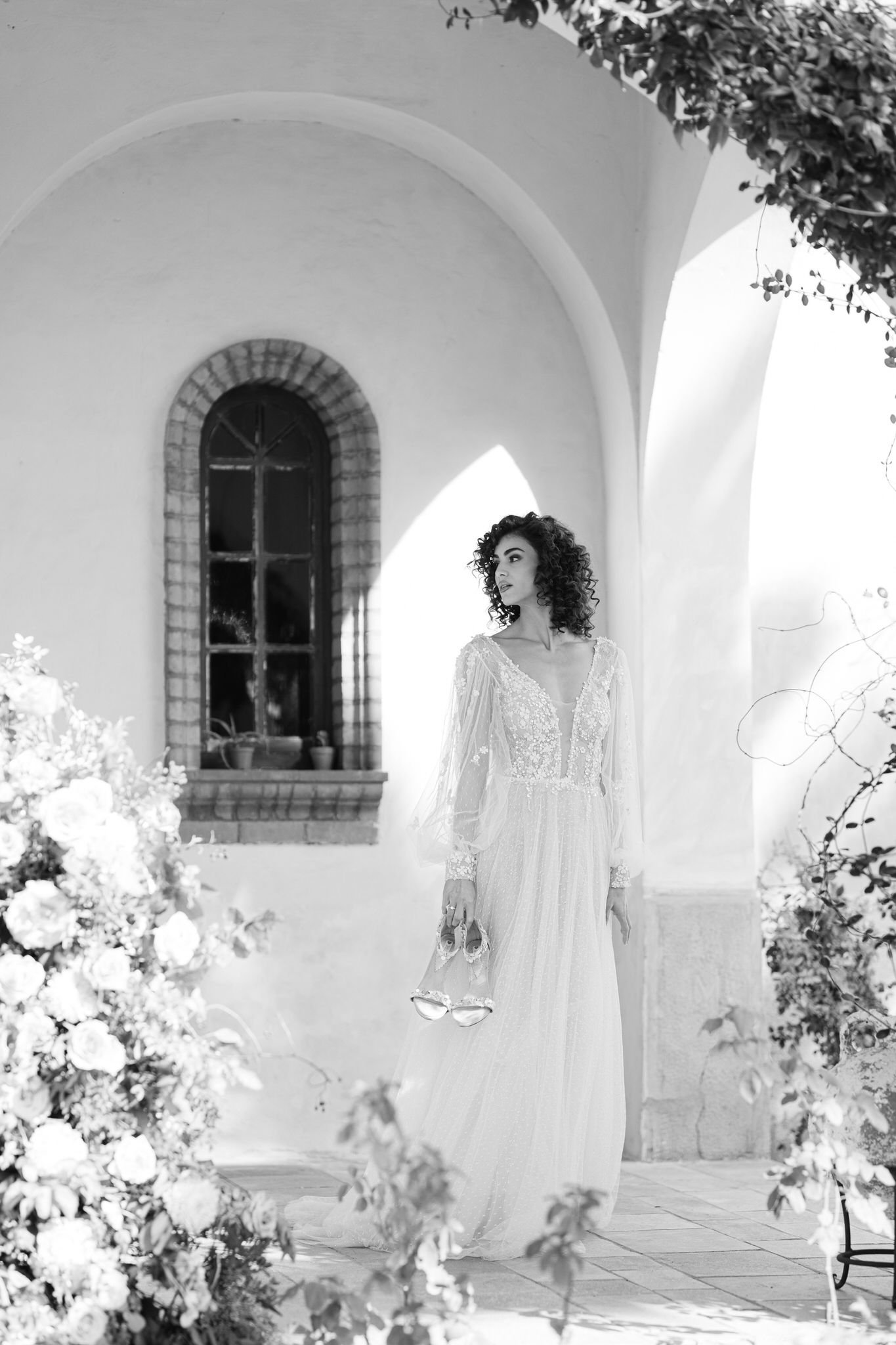 A romantic black and white photograph of a bride in a lace wedding gown standing outside a church with an arched window.