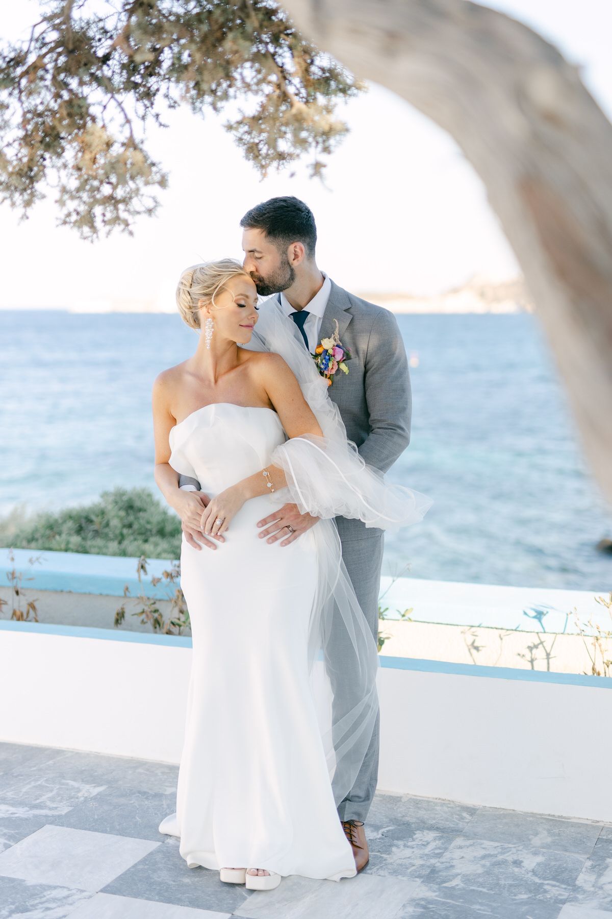 Couple poses romantically under an arched tree branch overlooking a Mediterranean coastal view on a sunny day.