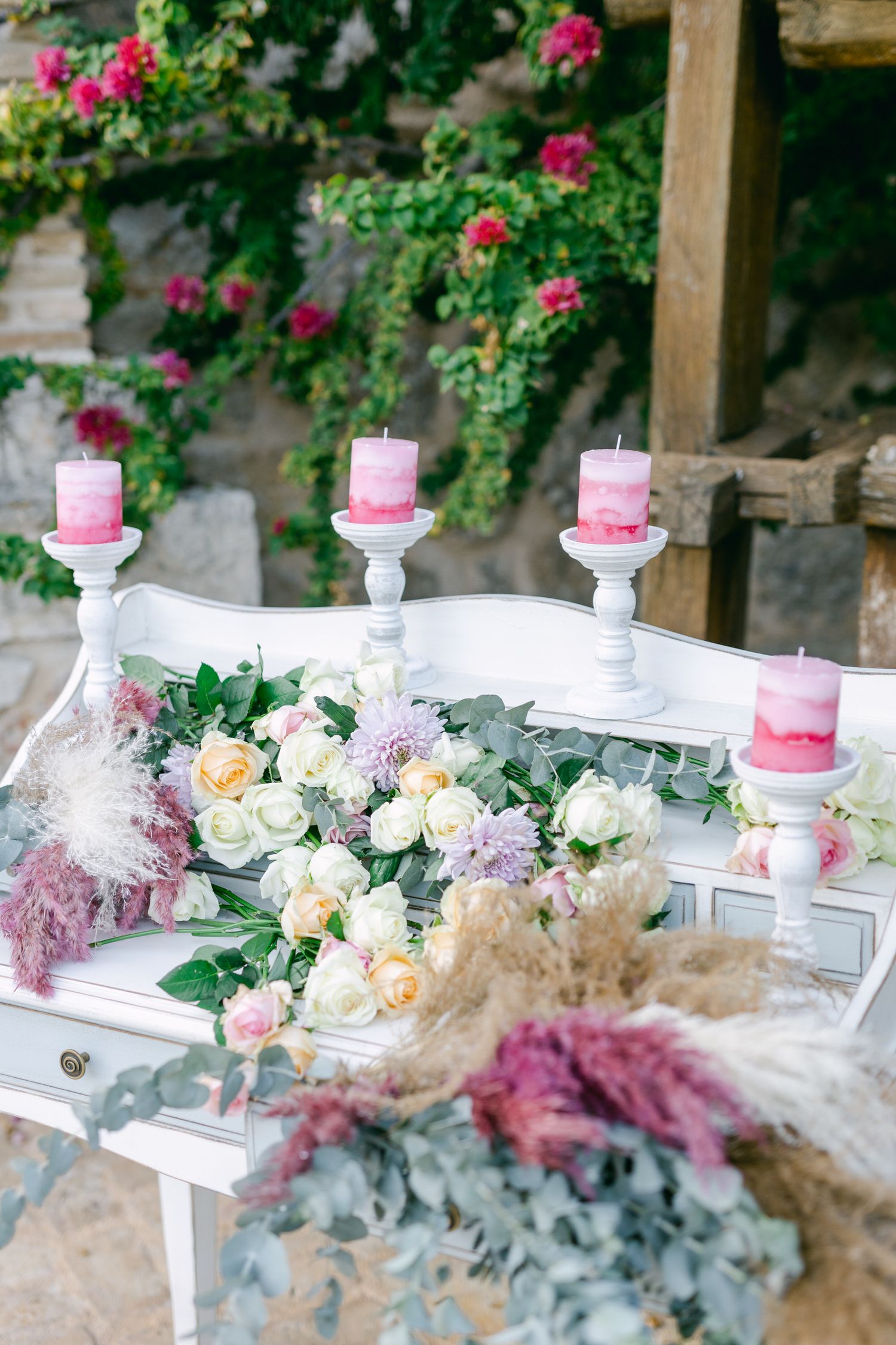 Romantic tablescape with pink candles, white roses, and dried flowers on vintage white furniture.