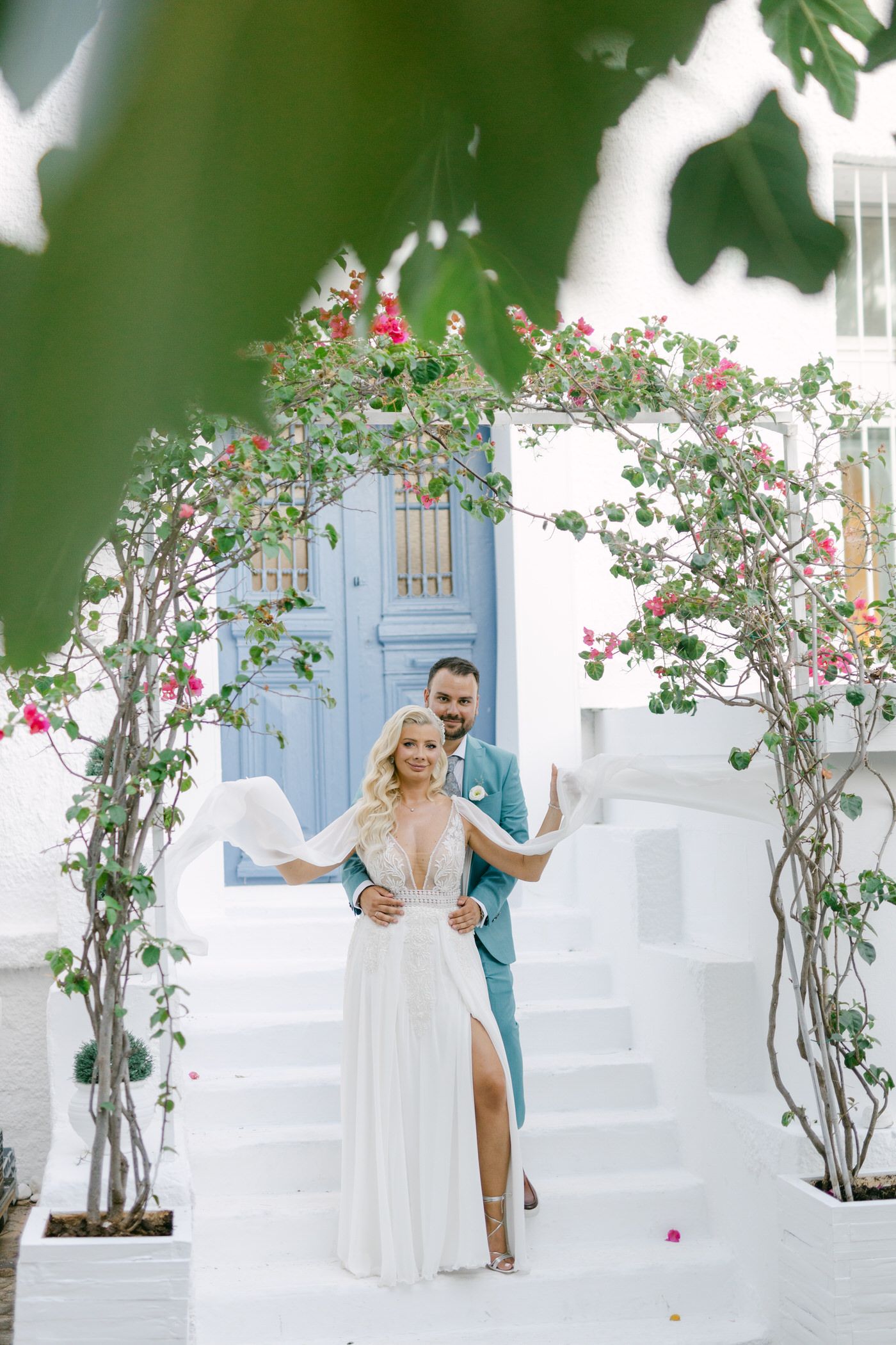Romantic wedding ceremony under a floral arch with climbing roses against white Mediterranean steps and blue door.