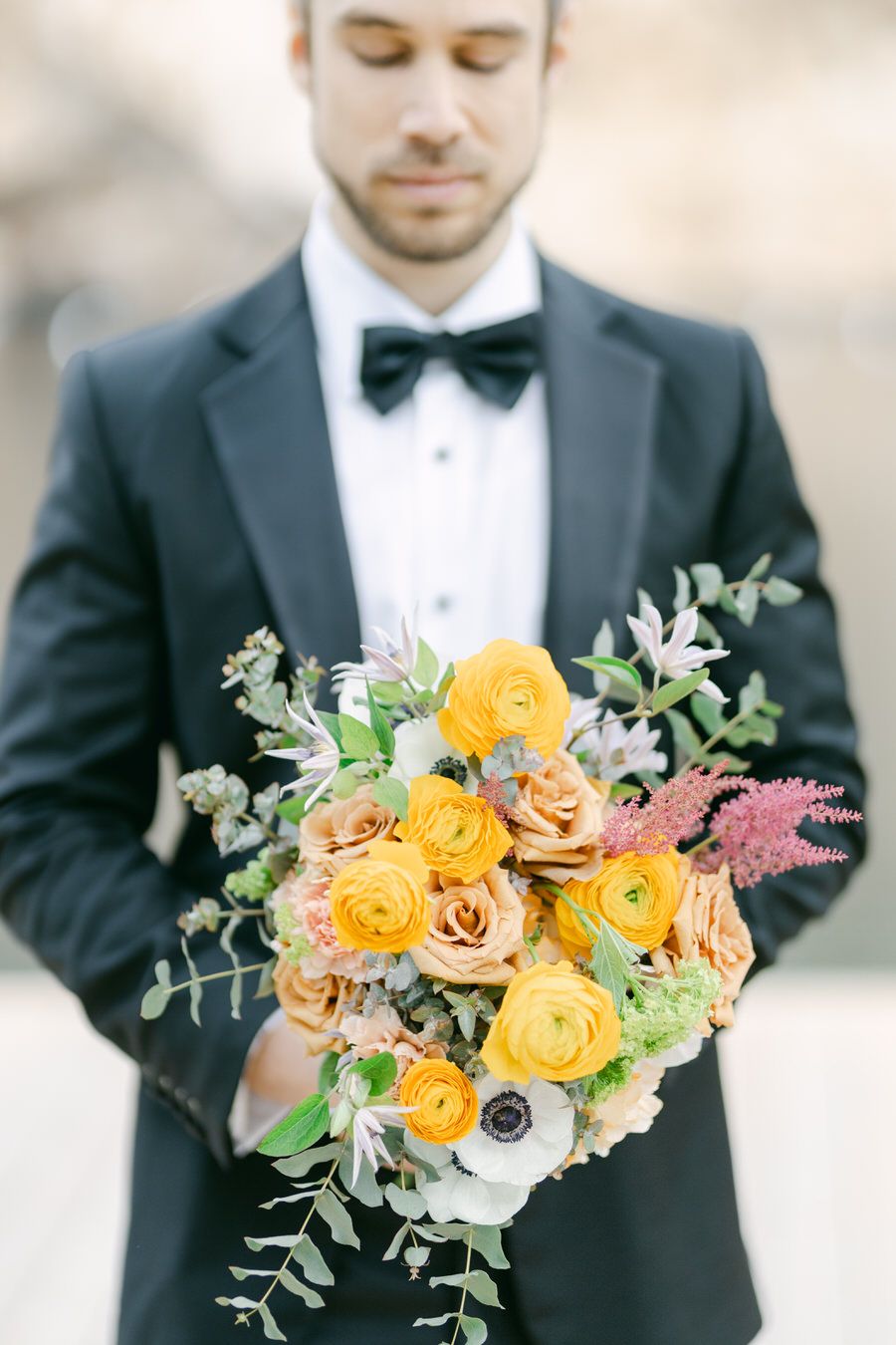 Wedding bouquet with bright yellow roses and eucalyptus held against black tuxedo.