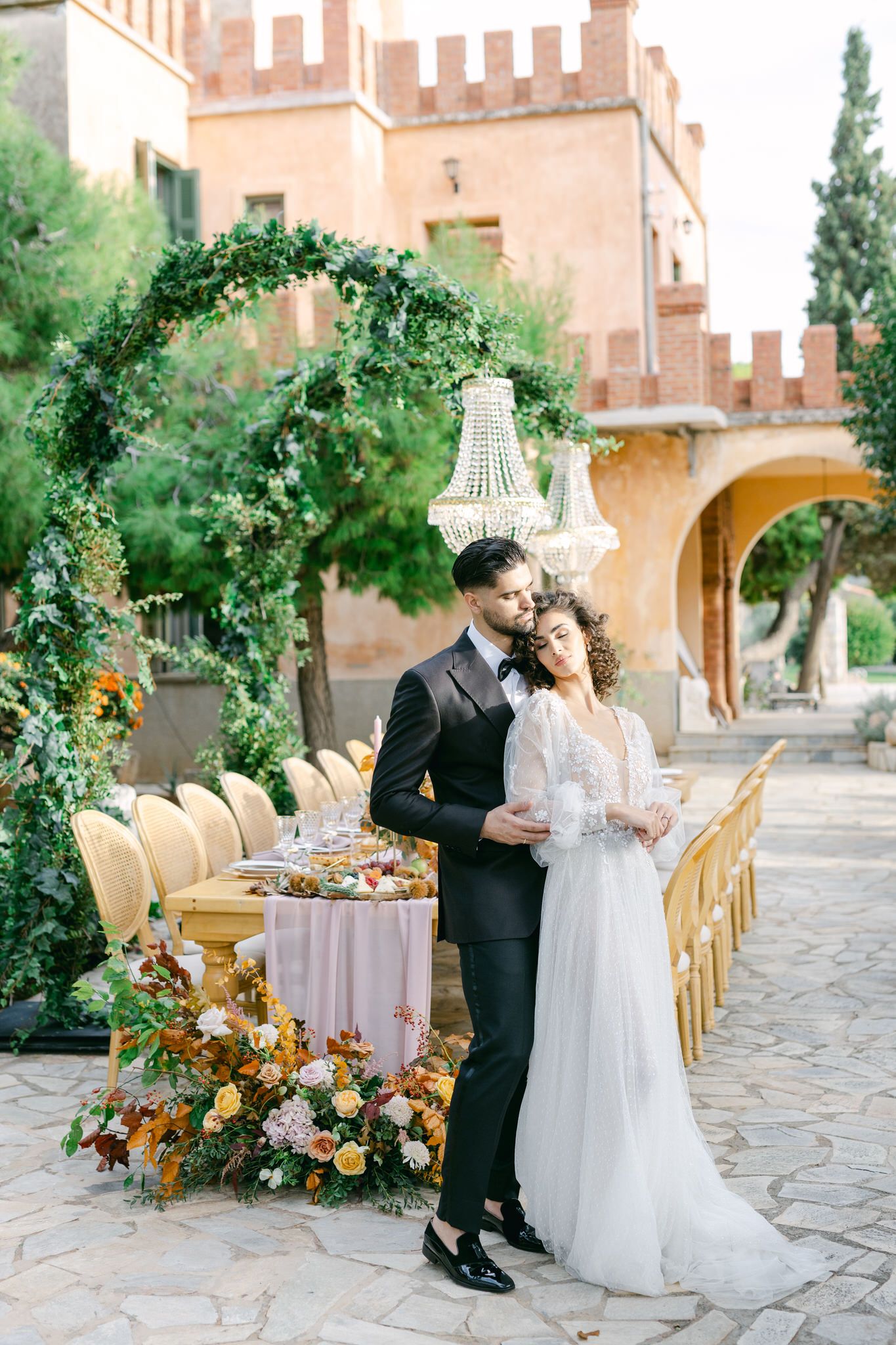Elegant wedding reception setup with floral arch and chandelier in front of rustic Mediterranean villa courtyard.