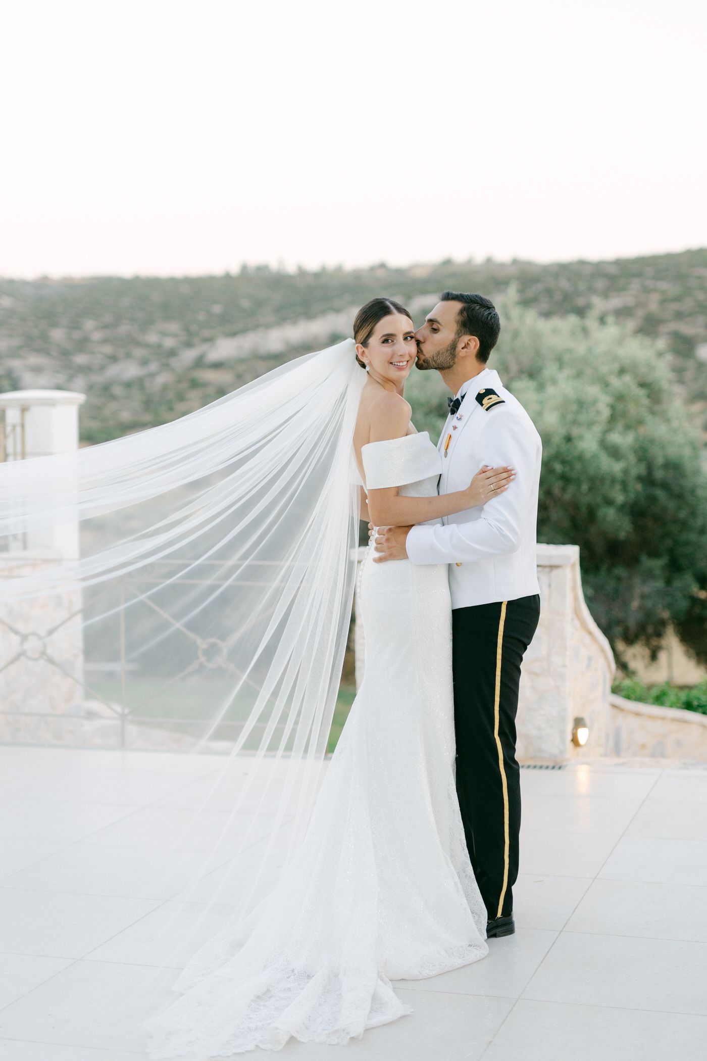 A romantic wedding couple embraces on a terrace with a flowing white veil and elegant formal attire.