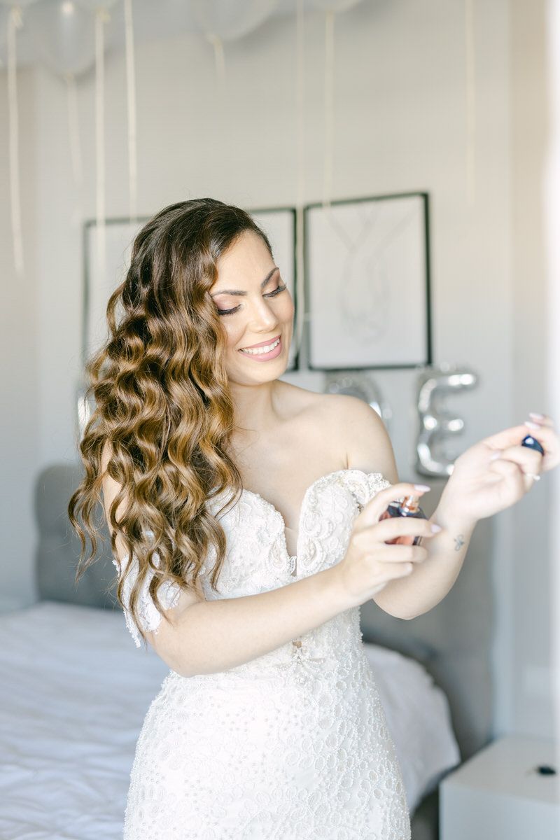A bride in a white lace gown with long wavy hair smiles while looking at her phone in a bedroom.