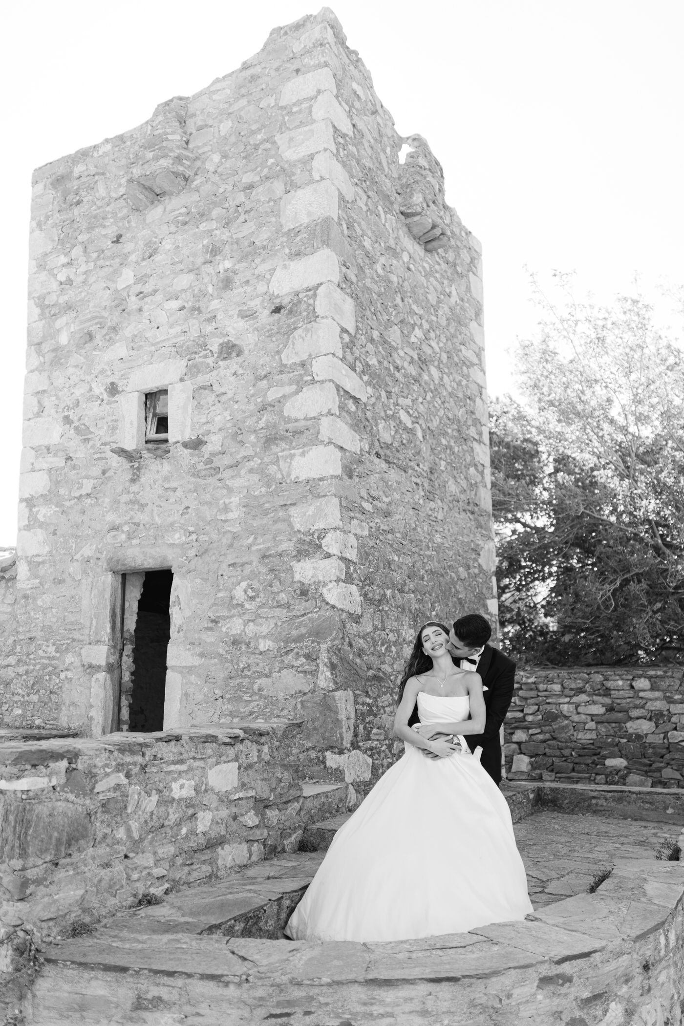 A couple in wedding attire stands by a stone tower structure in a black and white romantic photograph.