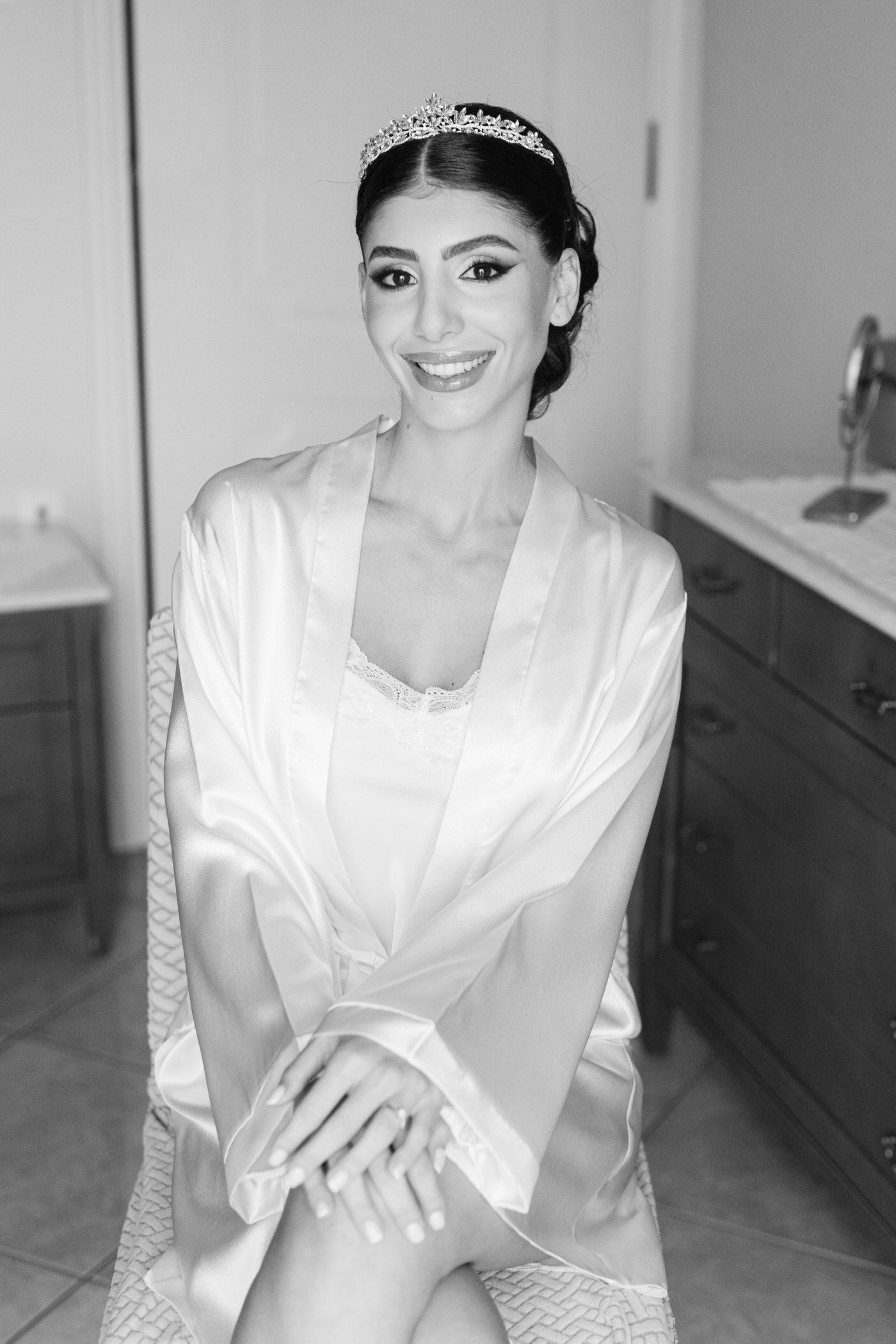 Beautiful black and white portrait of a bride in a silk robe and tiara smiling at the camera during wedding preparations.