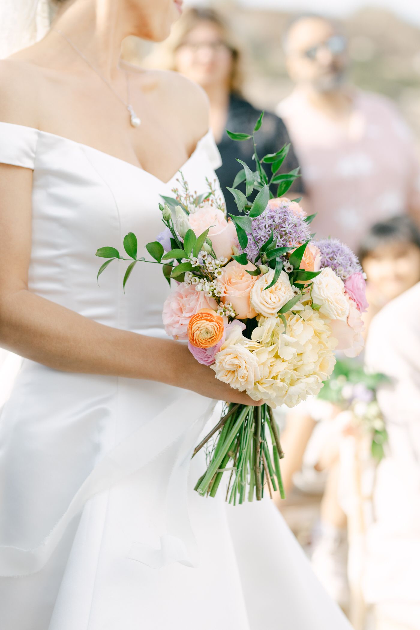 A delicate wedding bouquet with peach roses, white hydrangeas and lilac flowers held against a white bridal gown.