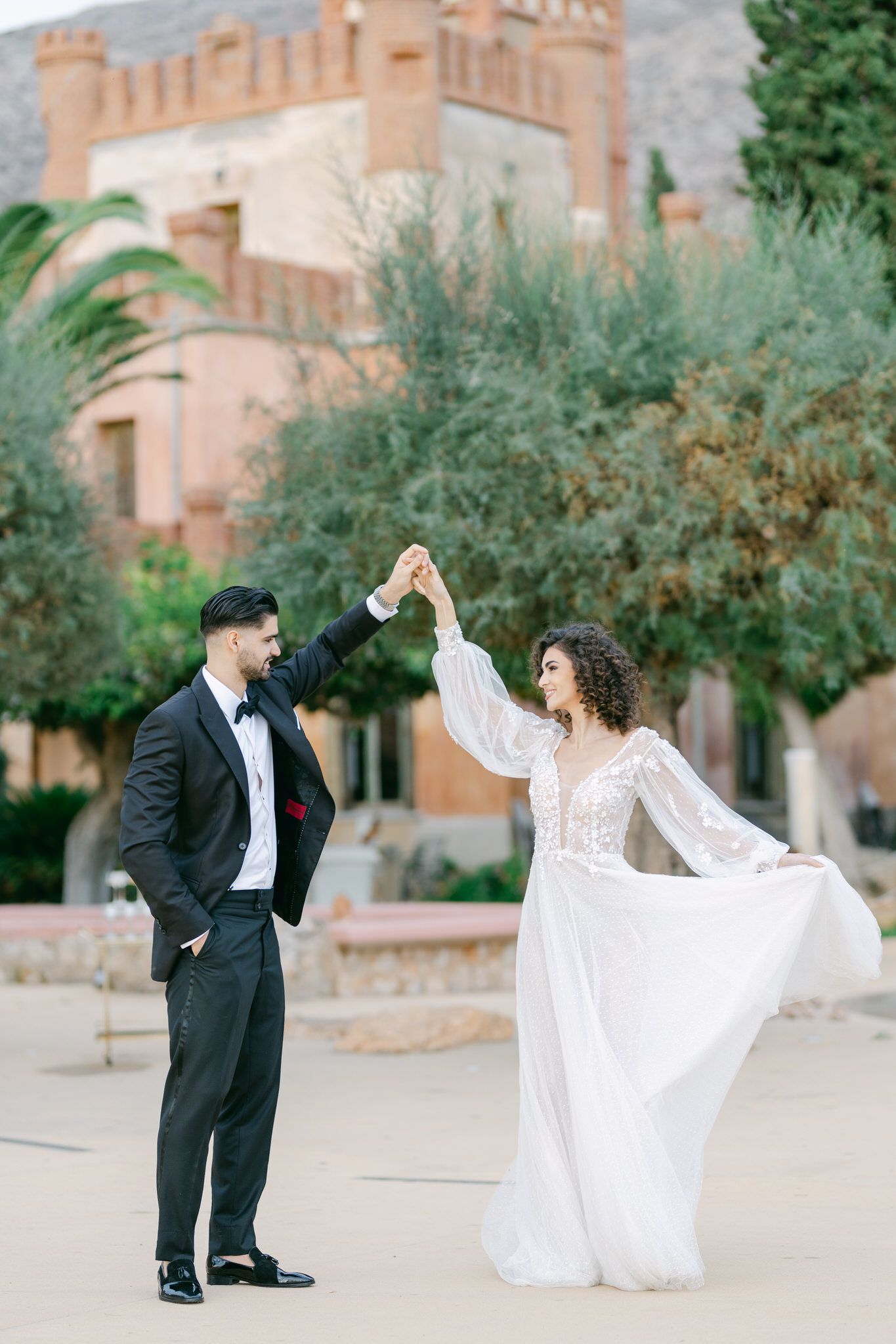 A couple in formal wedding attire dance together in front of a Mediterranean-style castle with lush gardens.