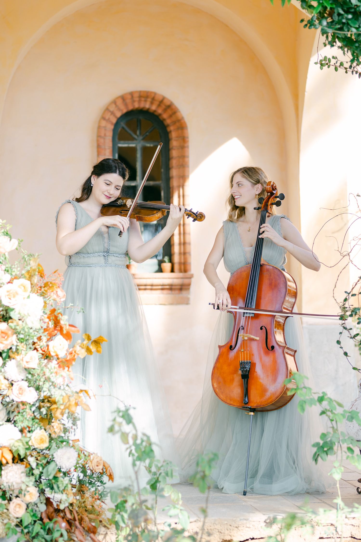 Musicians in soft blue dresses perform with violin and cello in front of an arched doorway with floral decorations.