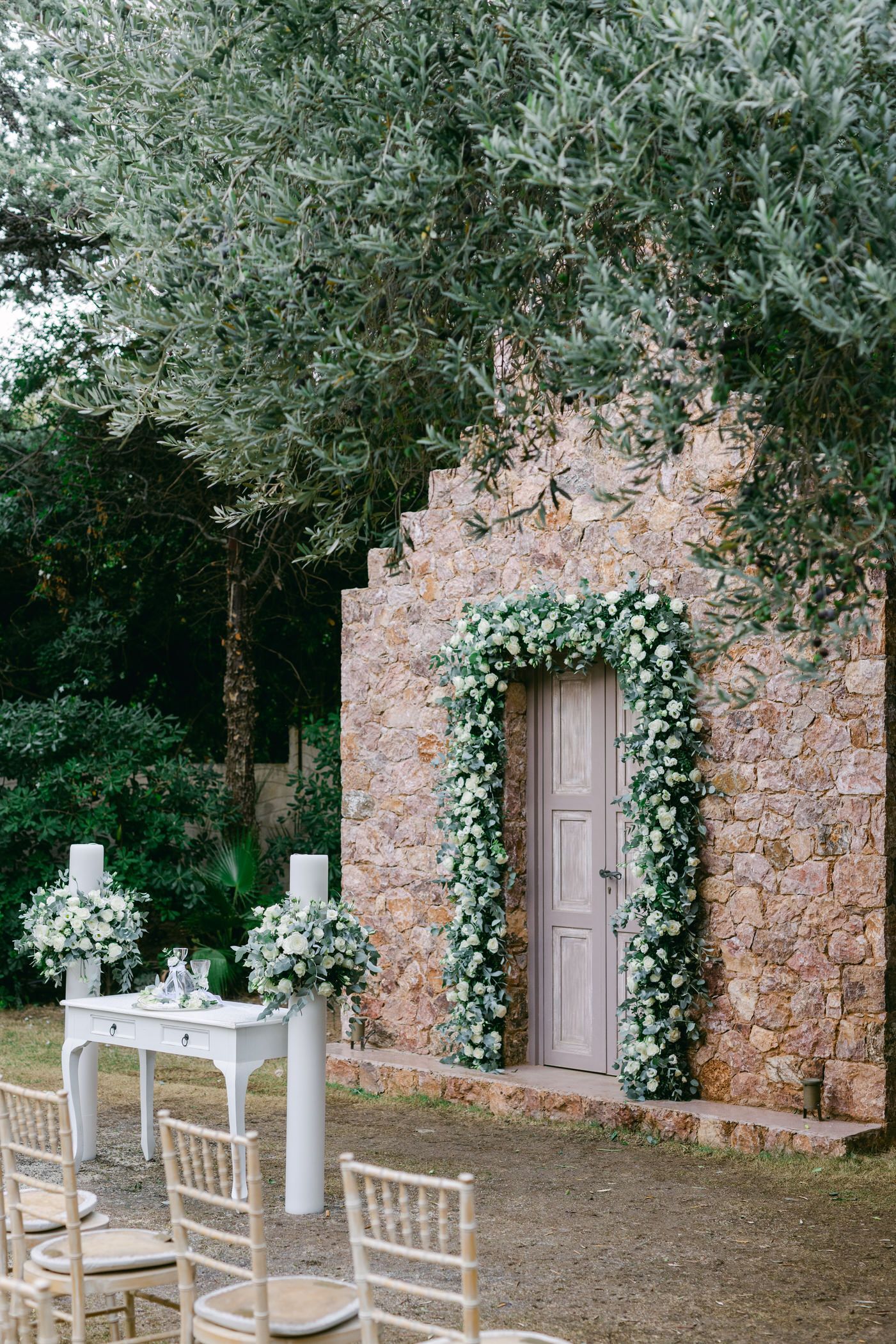 Romantic outdoor wedding setup with stone wall entrance adorned with greenery arch and white chiavari chairs.