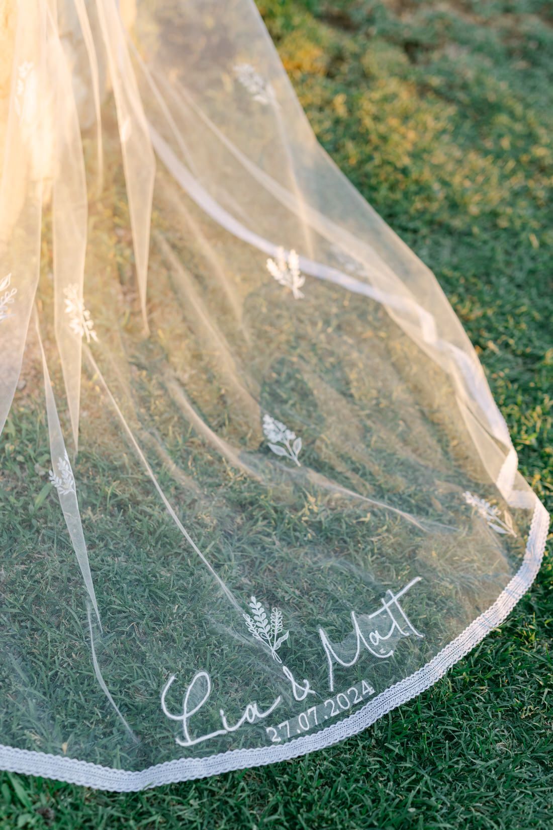 Close-up of delicate white wedding veil with embroidered lace details laying on green grass.