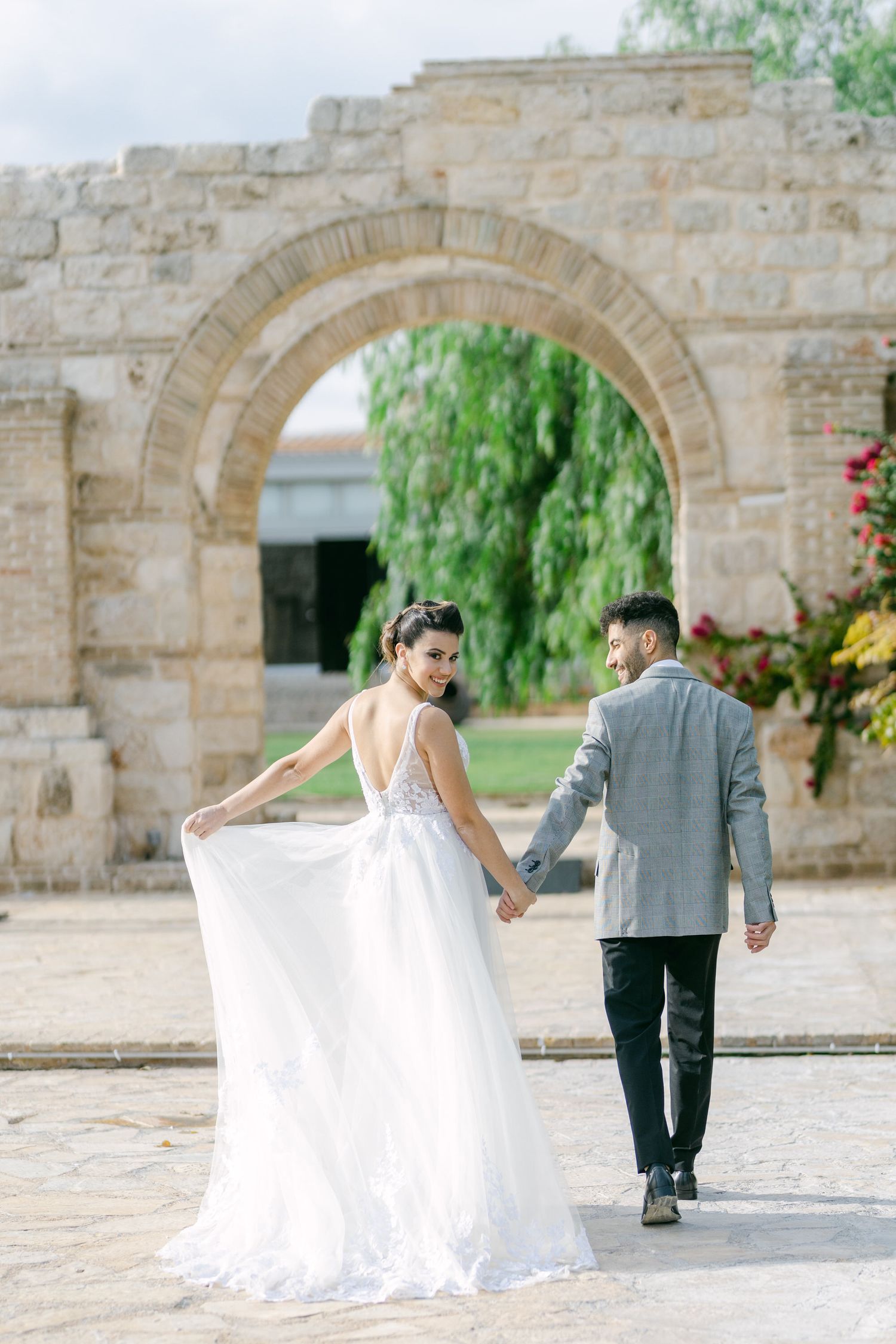 A romantic wedding couple walks through a stone archway surrounded by lush greenery and flowers.
