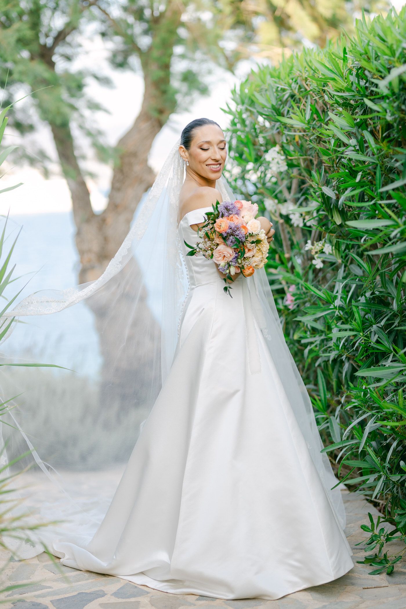 Elegant bride in flowing white wedding gown and veil holds vibrant bouquet while standing among lush garden foliage.