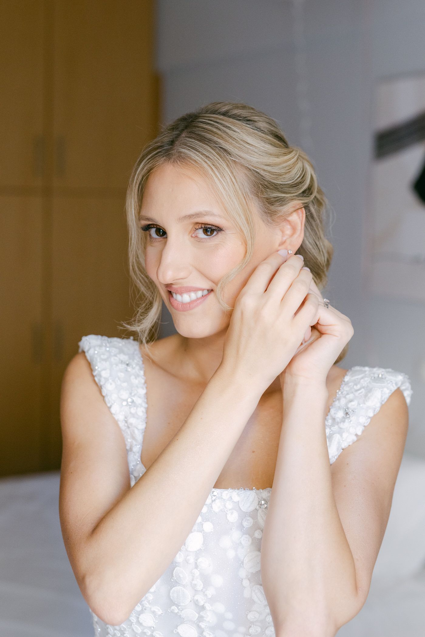 Elegant bride in white lace gown putting on earrings while smiling in brightly lit room.