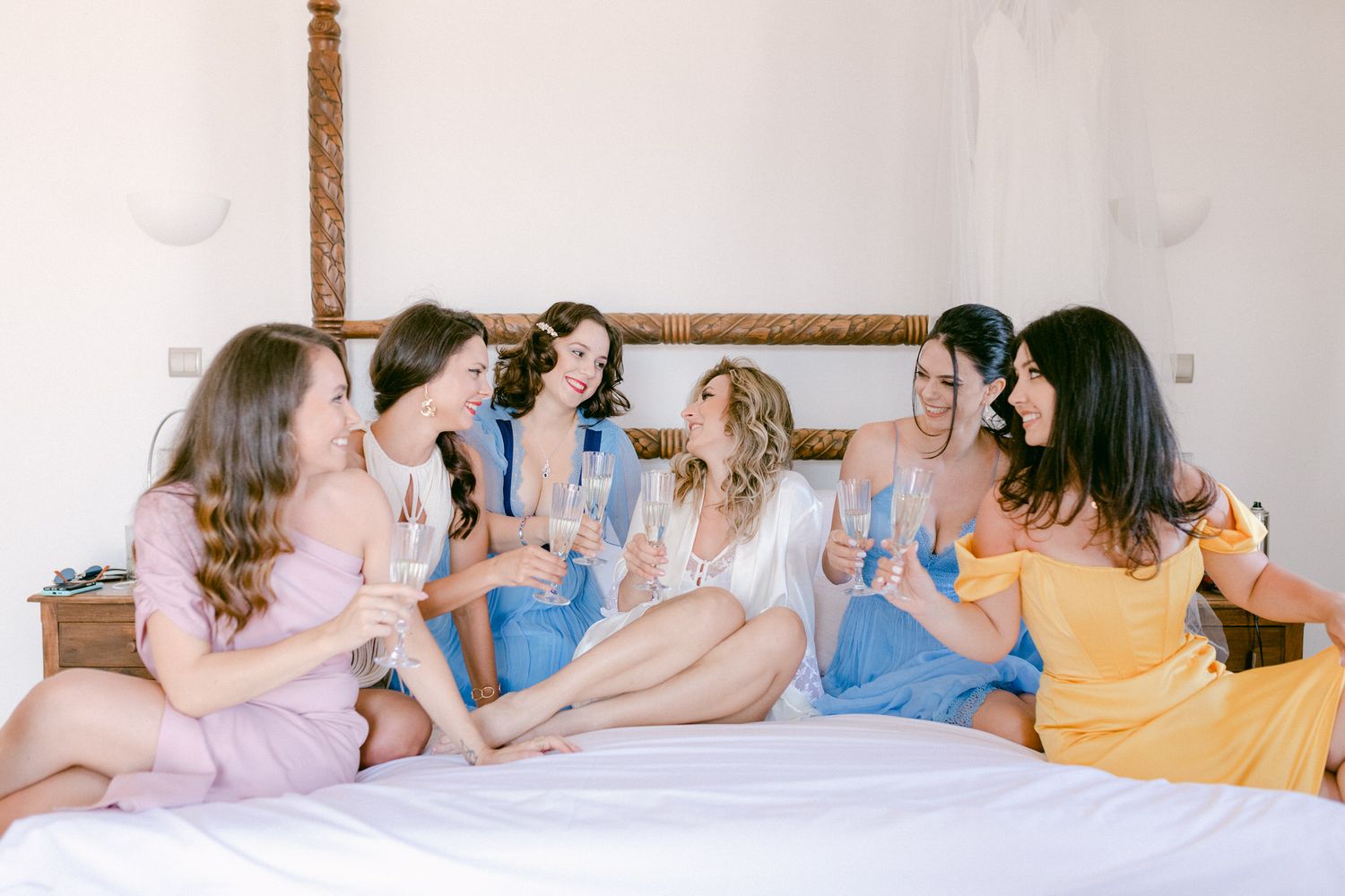 Group of friends in pastel dresses celebrating on a bed with drinks.