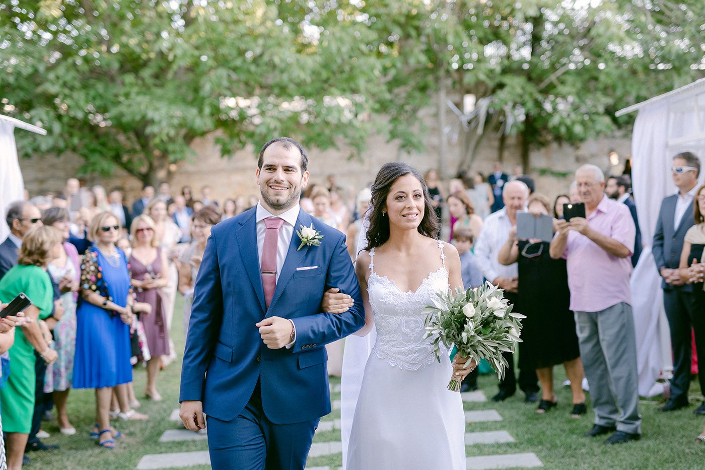 Two people walking down an outdoor wedding aisle while guests look on from either side in a garden setting.