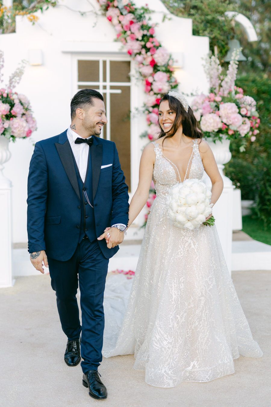 Wedding couple walking under floral arch decorated with pink roses and greenery.