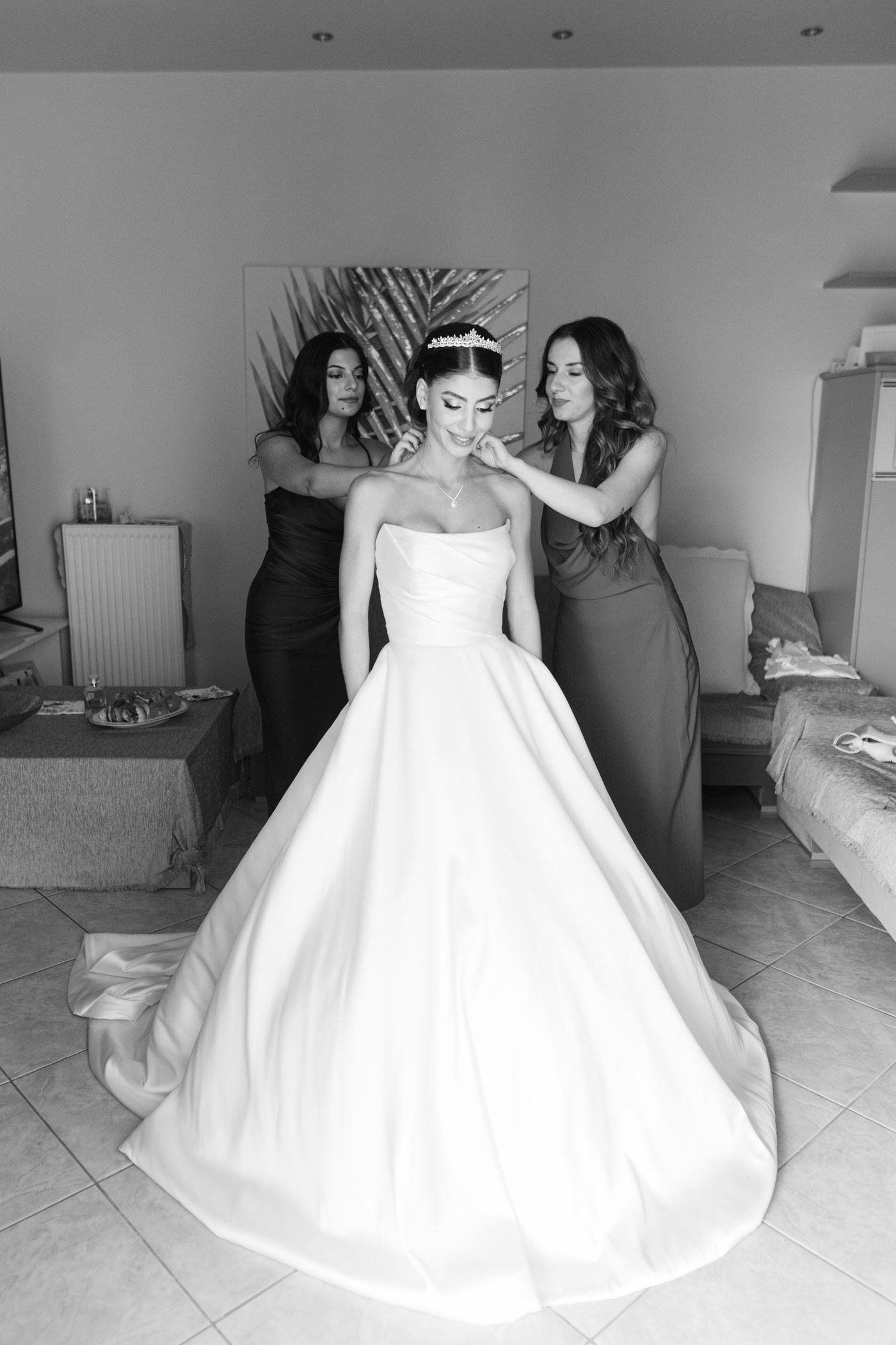 Black and white photo of wedding preparation scene with a bride in a classic ballgown being helped with final touches.
