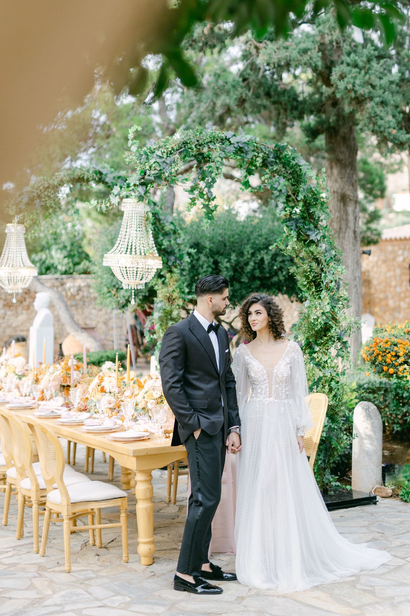 Elegant garden wedding reception setup with greenery arch, crystal chandeliers and golden table decorations outdoors.