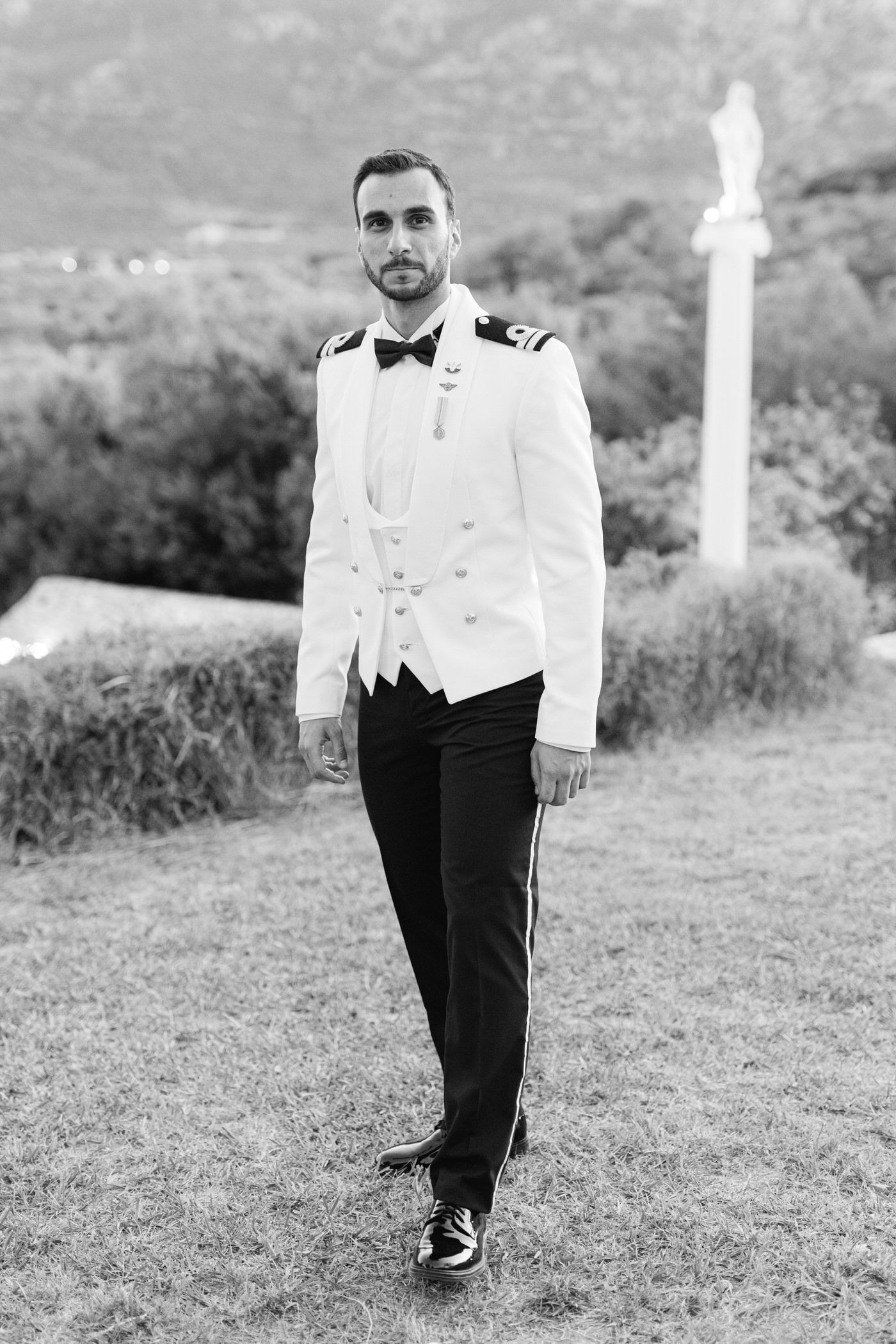 A formal black and white photo shows someone in an elegant white dinner jacket and bow tie standing outdoors.