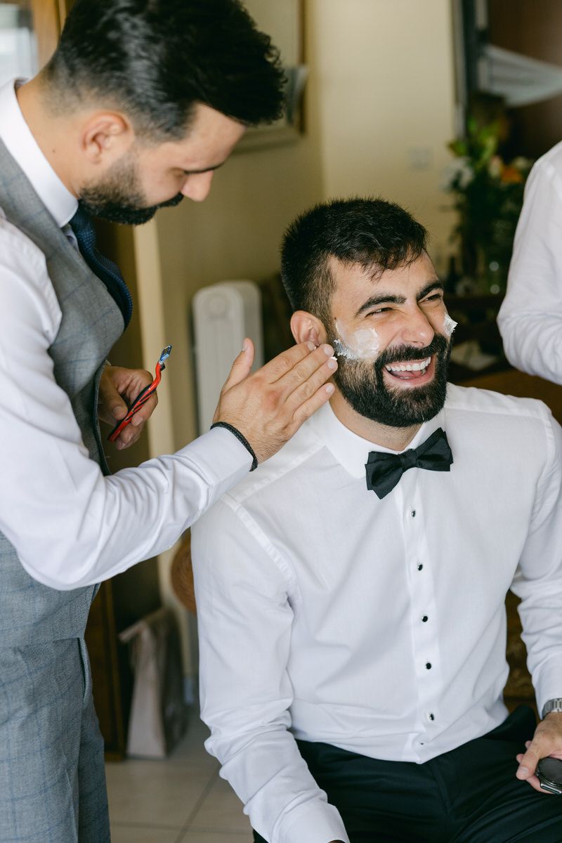 A groomsman in a gray vest helps with preparations while someone in a white dress shirt and bowtie smiles joyfully.