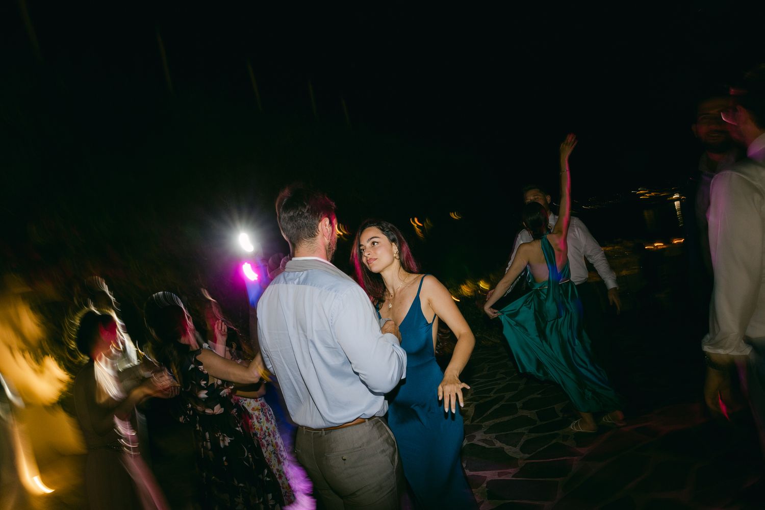 Couples dancing at a dimly lit evening reception with colorful lights and silhouettes in the background.