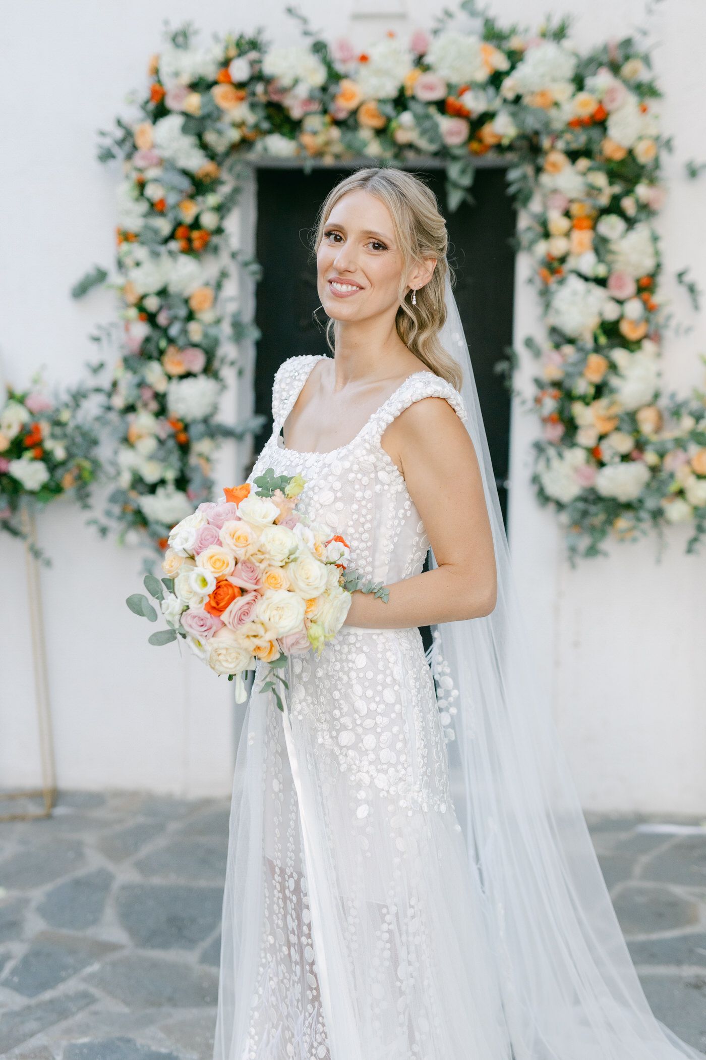 Bride in a delicate lace wedding dress holds a peach white bouquet under a floral arch with orange blooms. Κτήμα Αργιθέα Tonia and Theodore