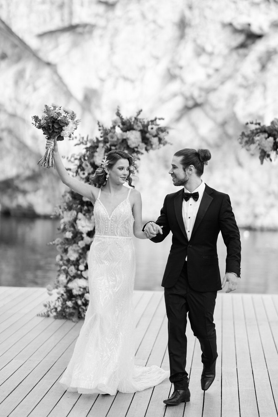 A black and white photo of a newlywed couple walking together on a dock.