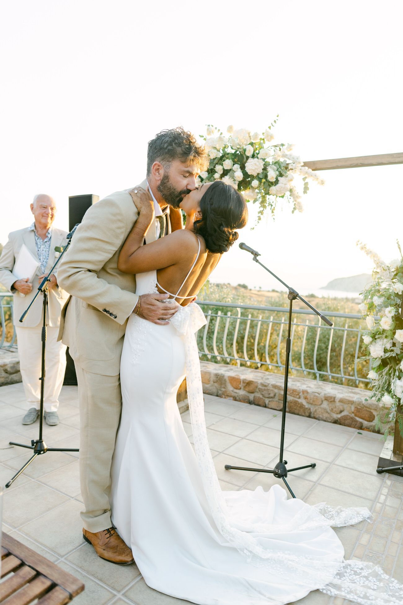 Newlyweds embrace during first kiss at outdoor ceremony with scenic backdrop.