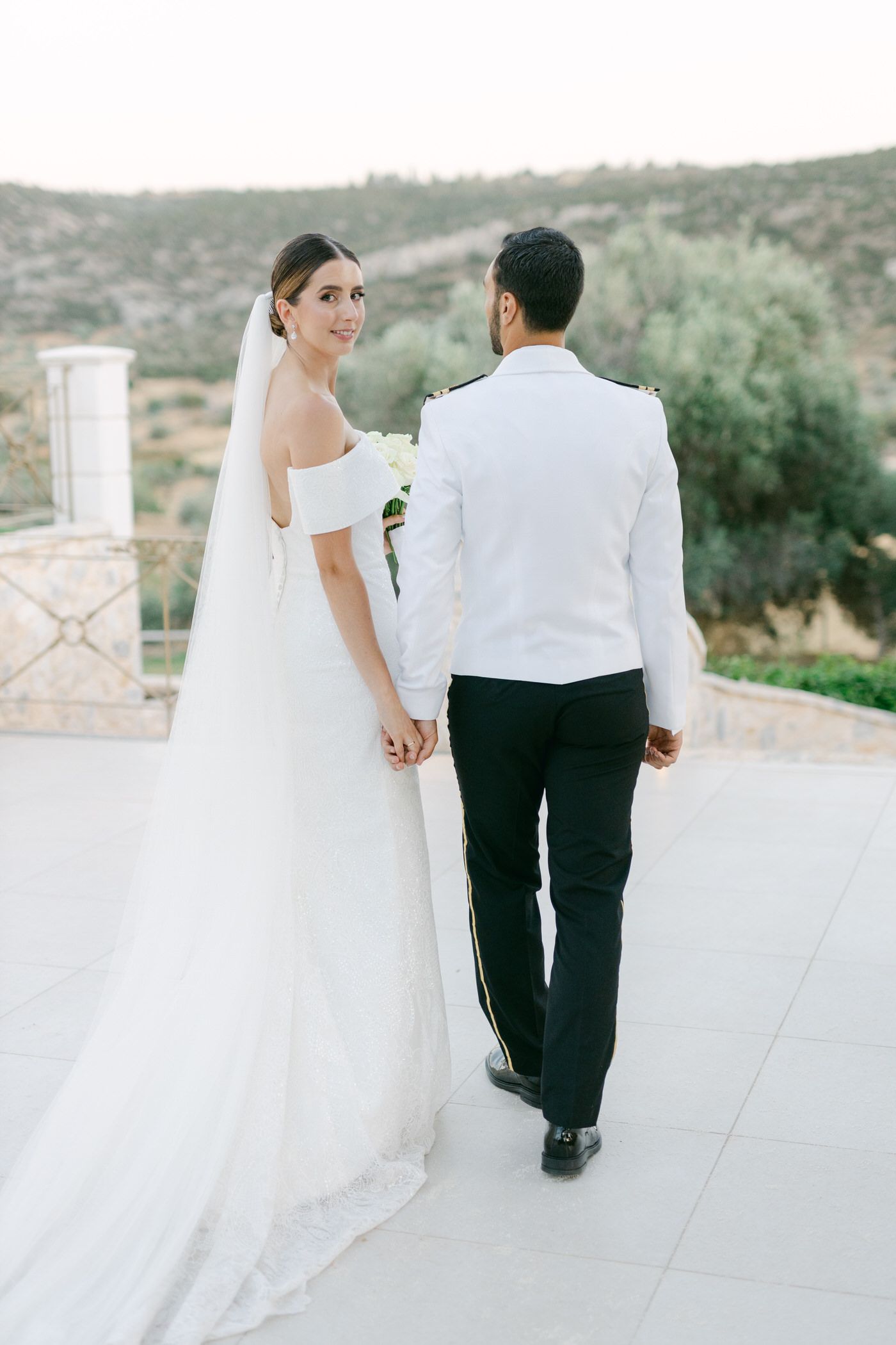 Newlywed couple in formal wedding attire holding hands while walking on an outdoor terrace with scenic mountain views.