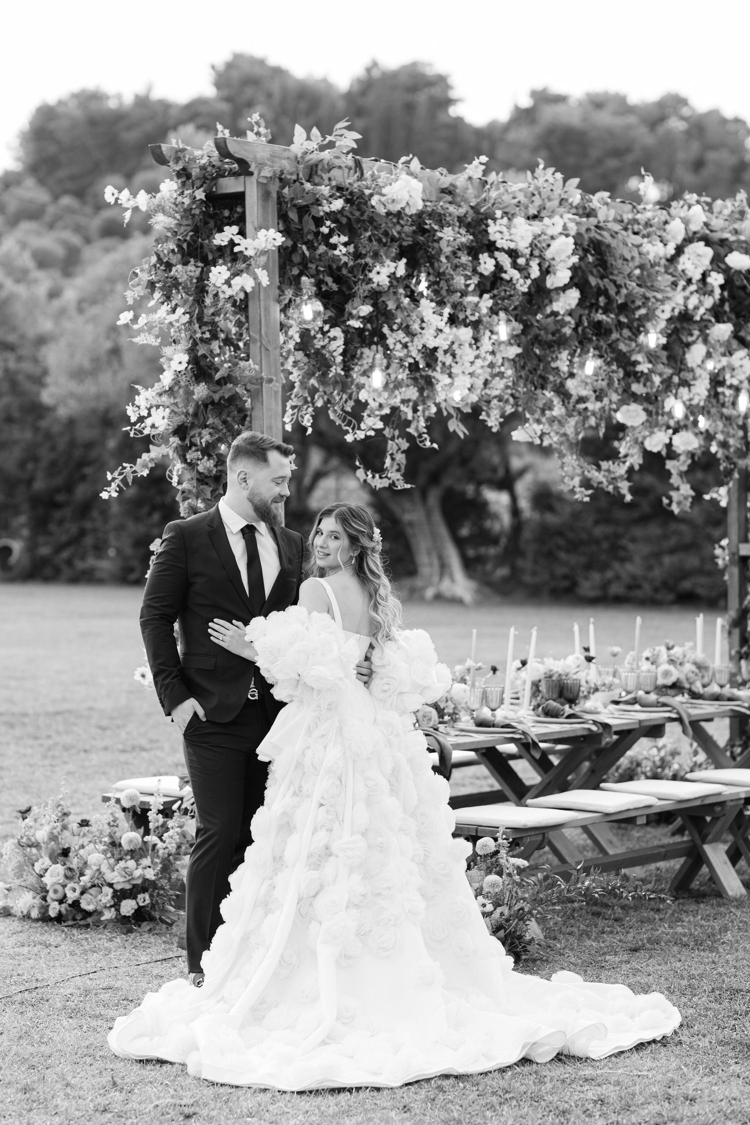 Black and white wedding photo showing couple embracing under floral arch by rustic outdoor table setting.