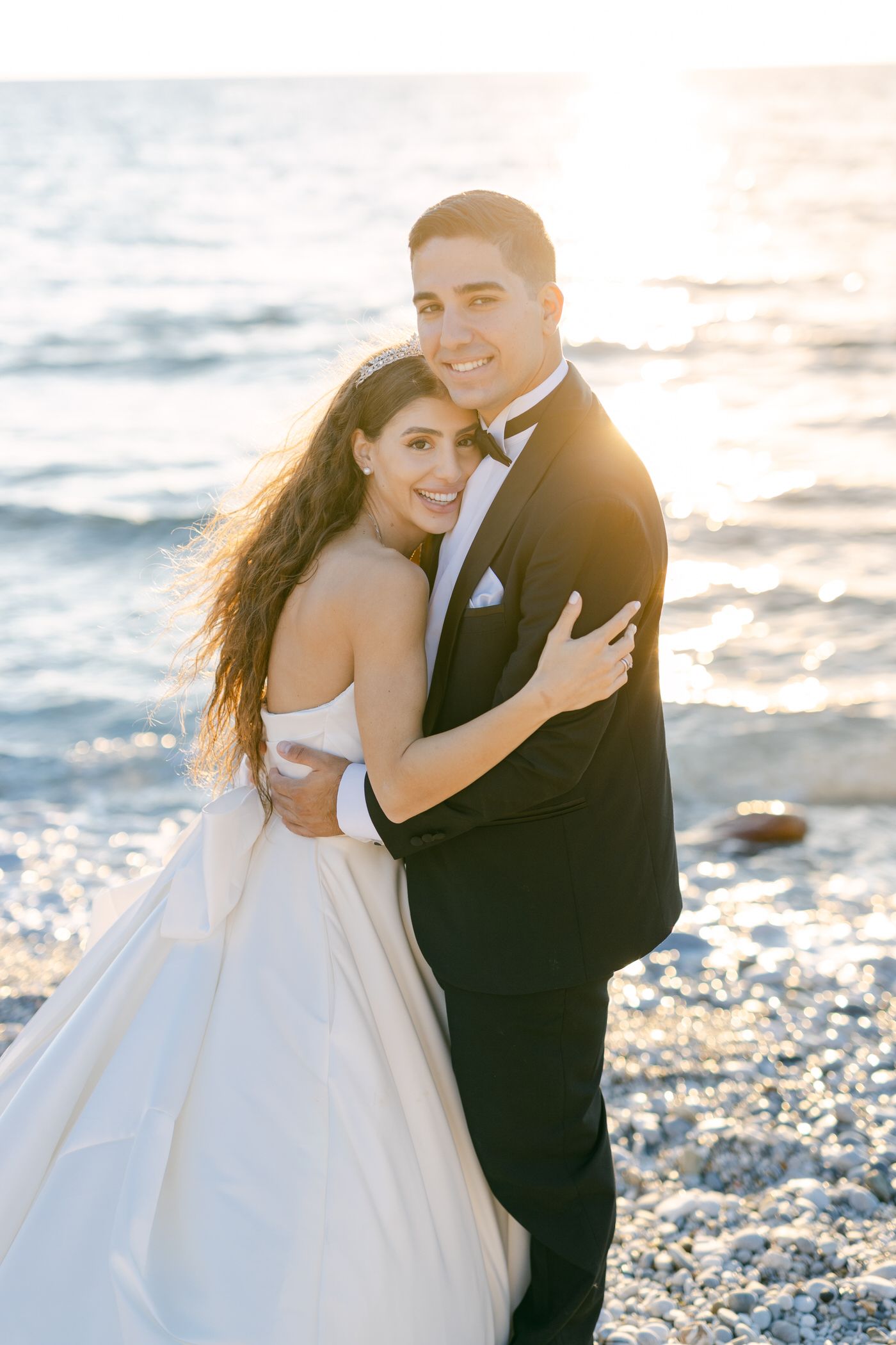 A romantic wedding couple embraces on a beach at sunset with waves in the background.
