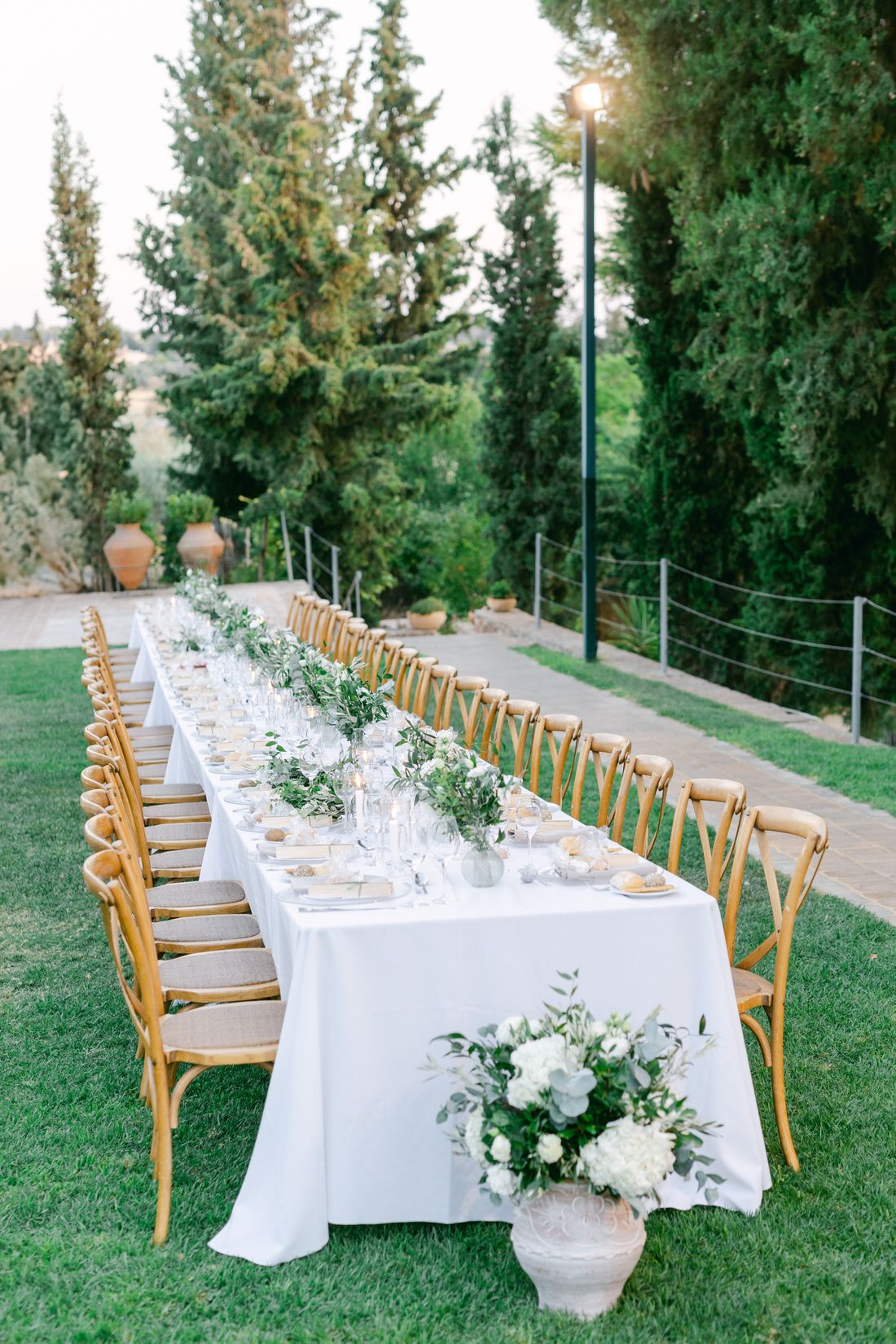 Elegant outdoor wedding table setting with white linens, wooden chairs, and natural greenery under tall cypress trees.