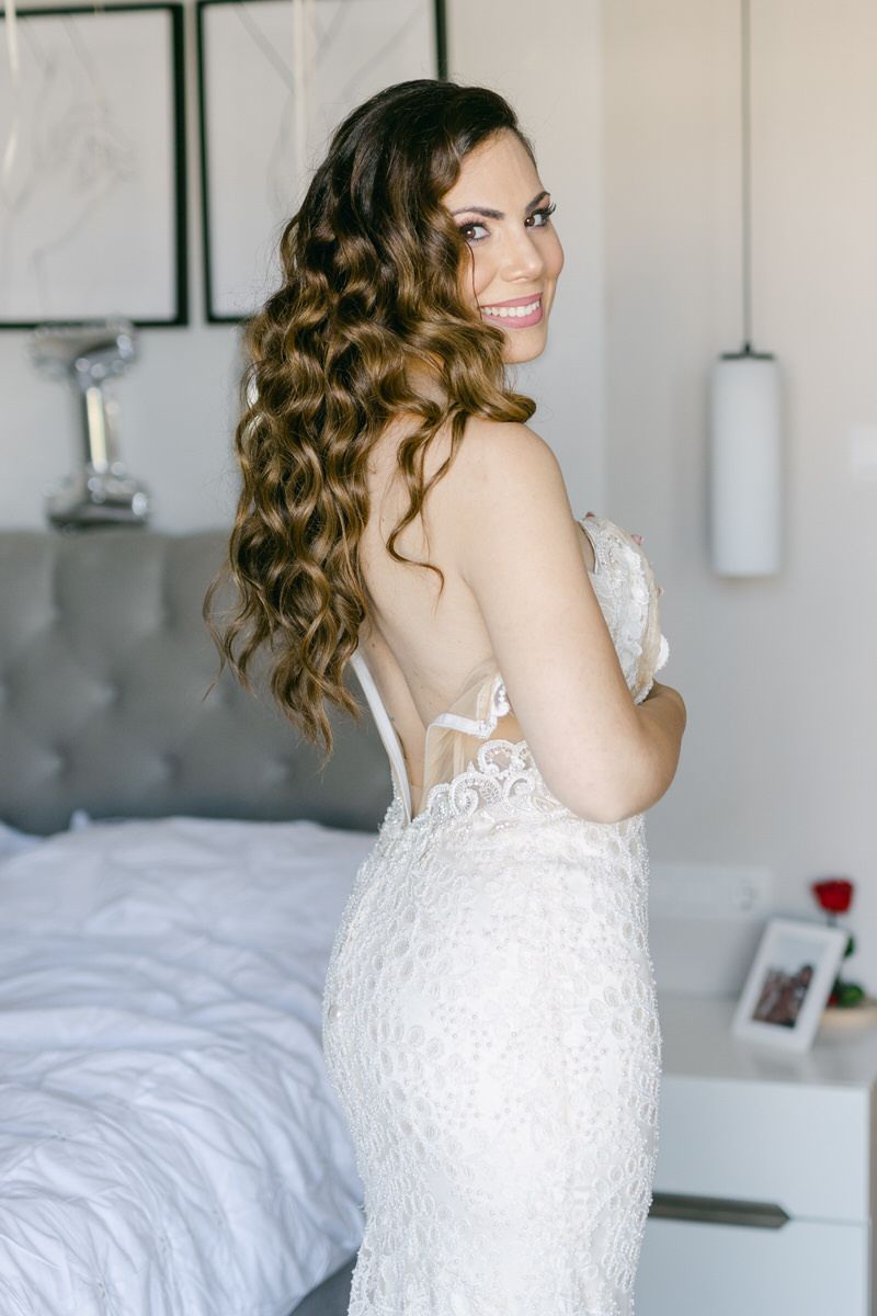 A bride in a fitted lace wedding dress poses in a bedroom, showing off long curled brown hair and elegant styling.