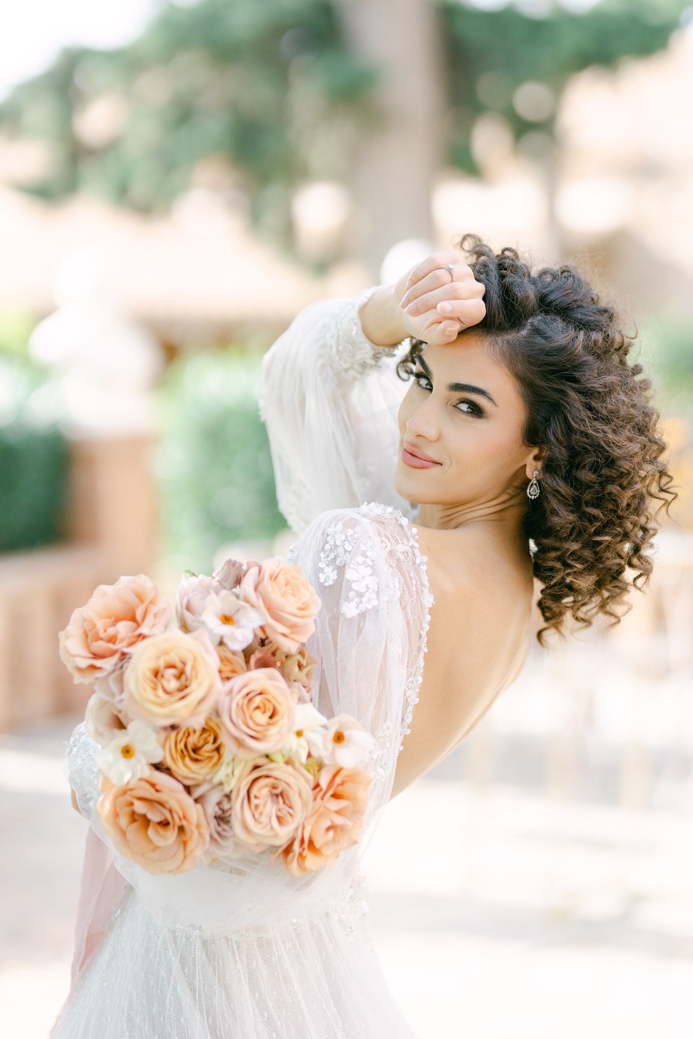 Romantic bridal portrait with peachy garden roses and a flowing lace gown against a soft blurred outdoor background.