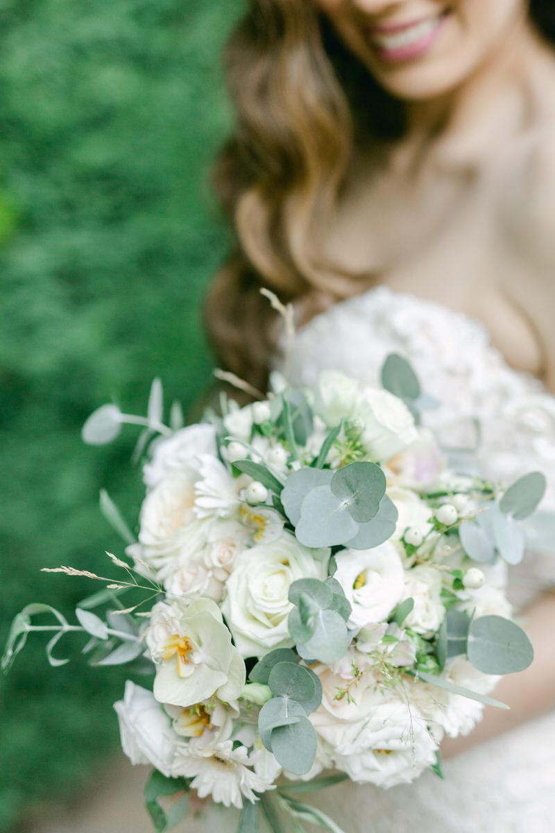 A romantic white wedding bouquet with roses, eucalyptus leaves and delicate floral arrangements against a green background.