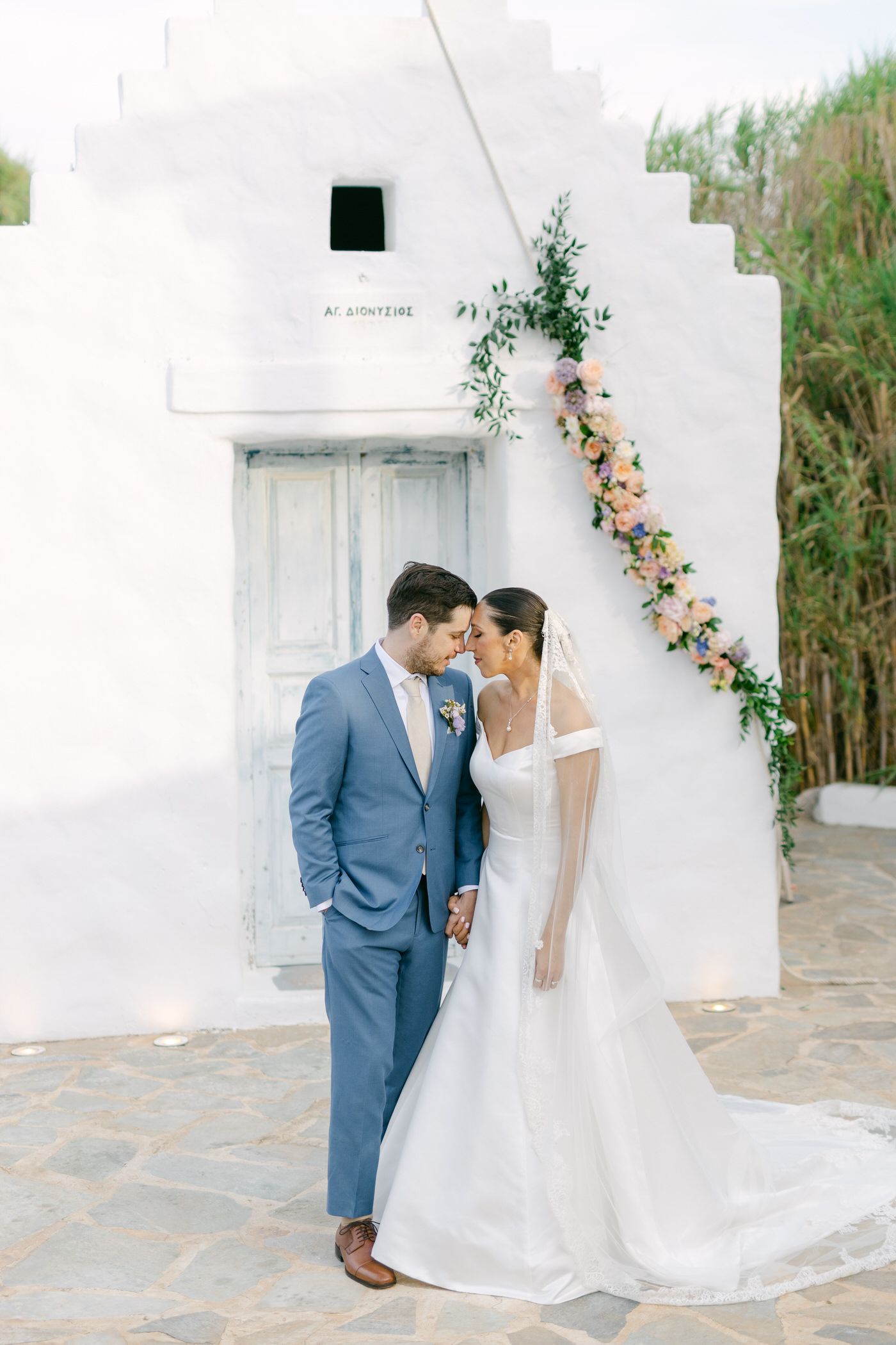 A wedding couple stands intimately in front of a white Mediterranean-style building with floral garland decor.