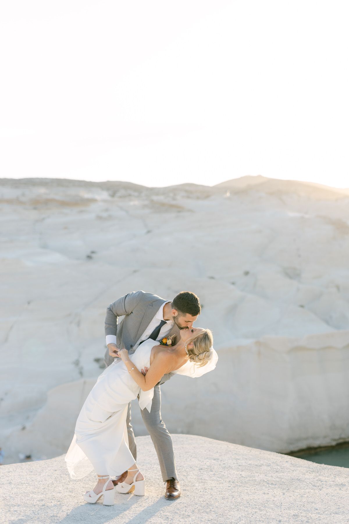A couple in wedding attire shares a romantic dip pose against a white rocky desert landscape at sunset.
