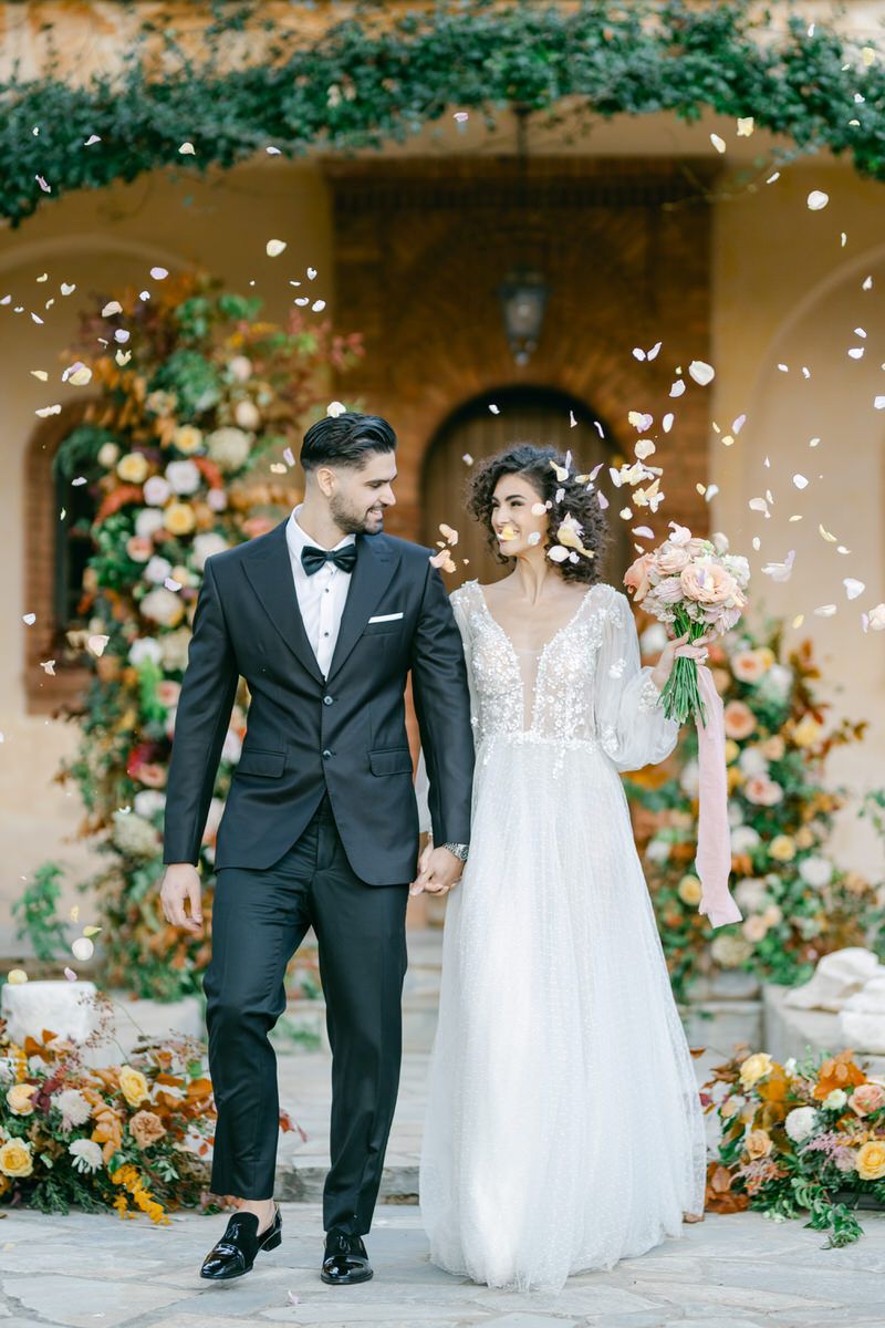 Wedding couple walking hand in hand along stone pathway with flower petals falling and floral decorations.