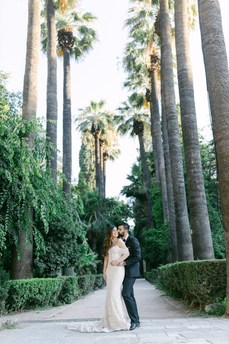 A couple embraces on a garden path lined with towering palm trees and manicured hedges.