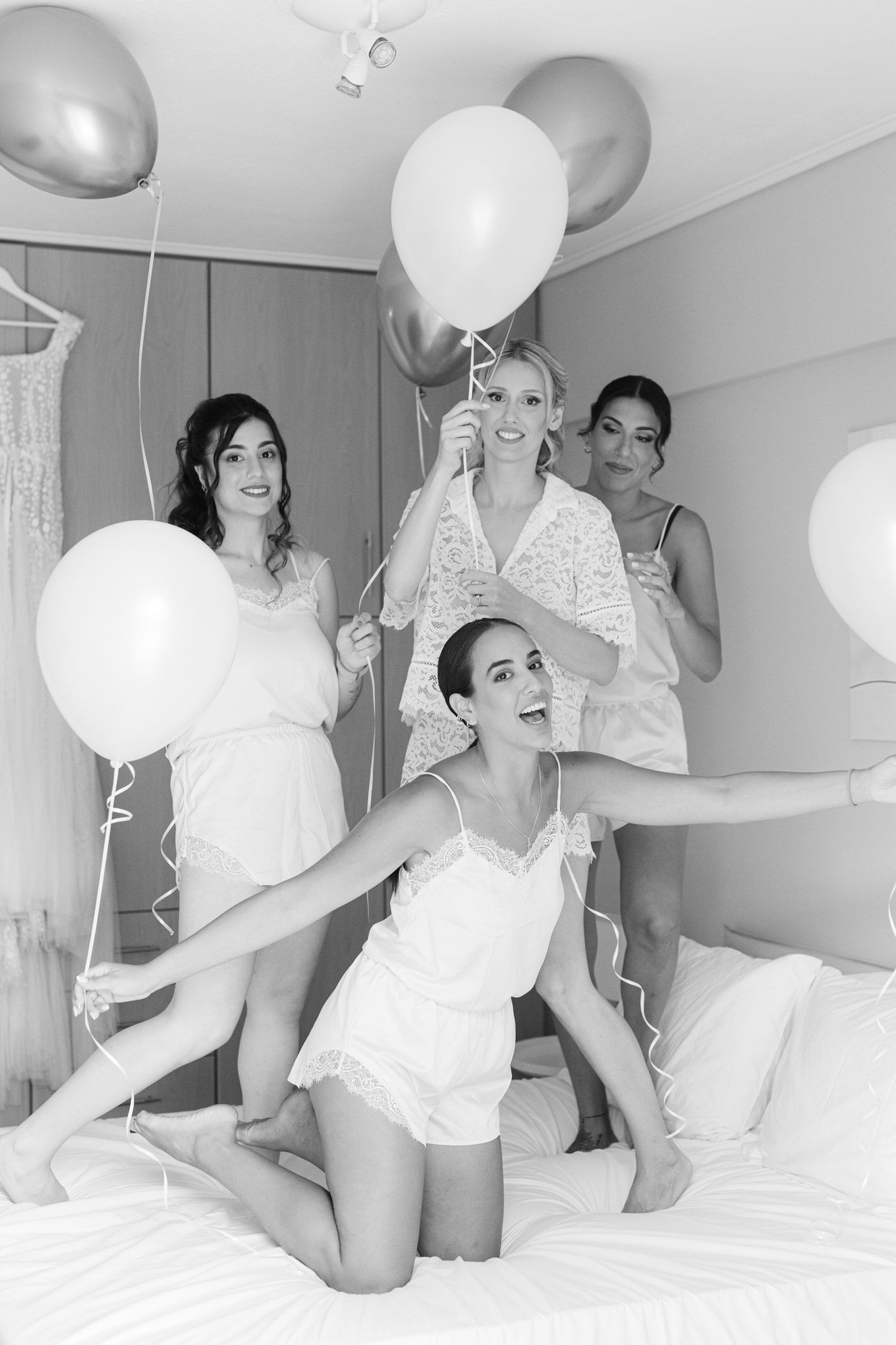 Black and white photo of a bridal party in white outfits having fun with balloons on a bed during pre-wedding preparations.
