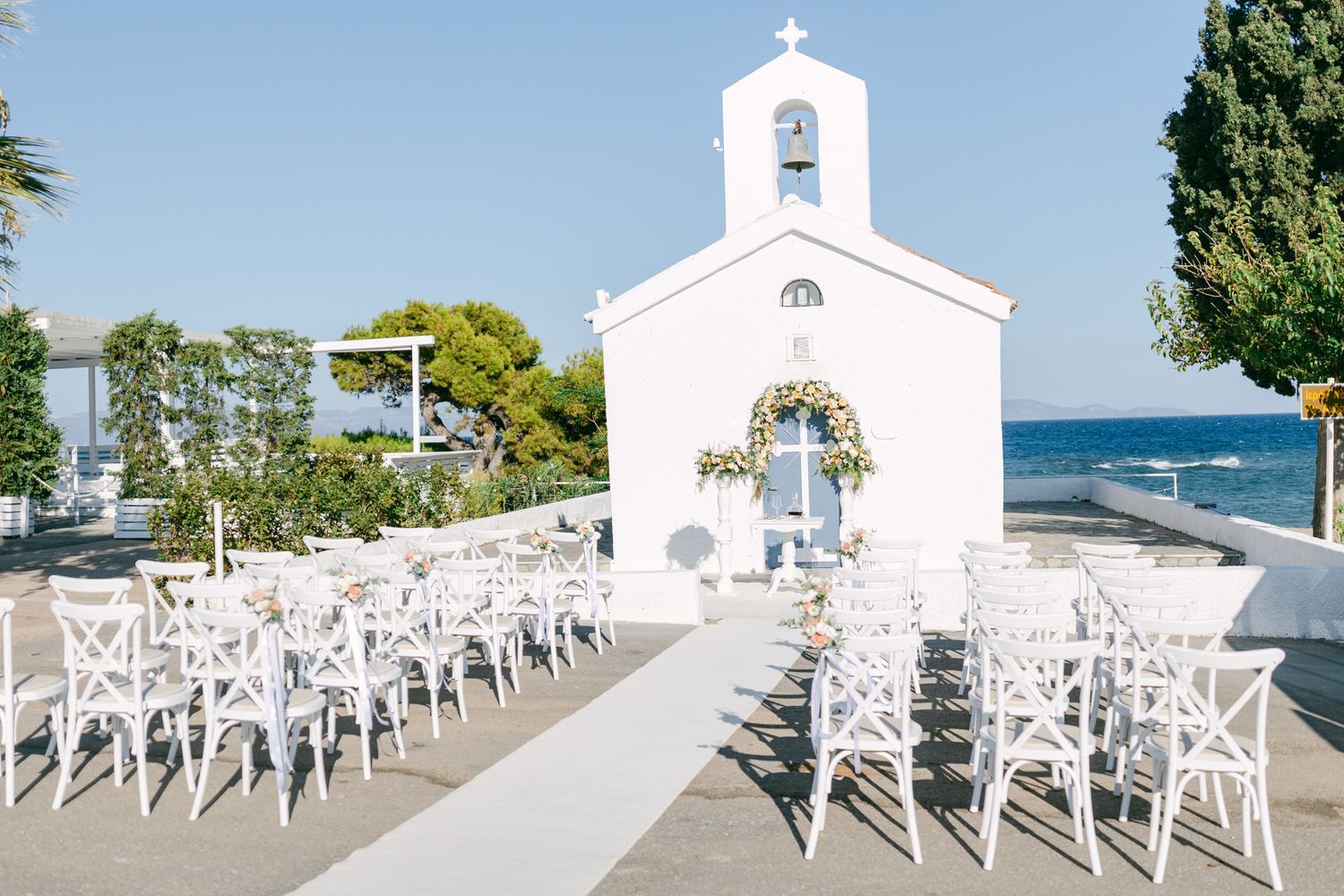 White chairs line up before a small seaside chapel with blue ocean views for an outdoor wedding ceremony.