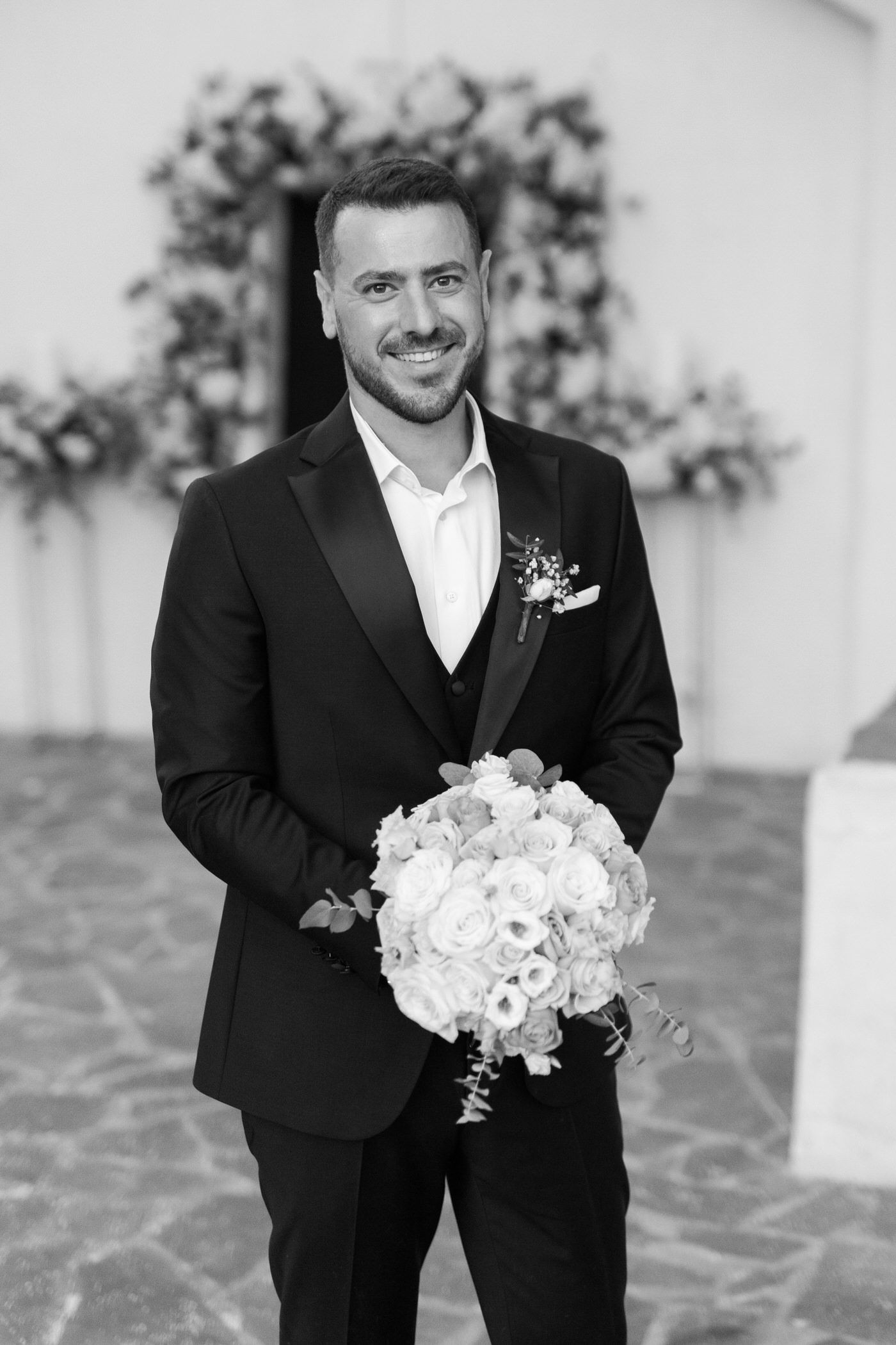 A groom in a classic black tuxedo holds a beautiful white floral bouquet while smiling at a wedding venue.