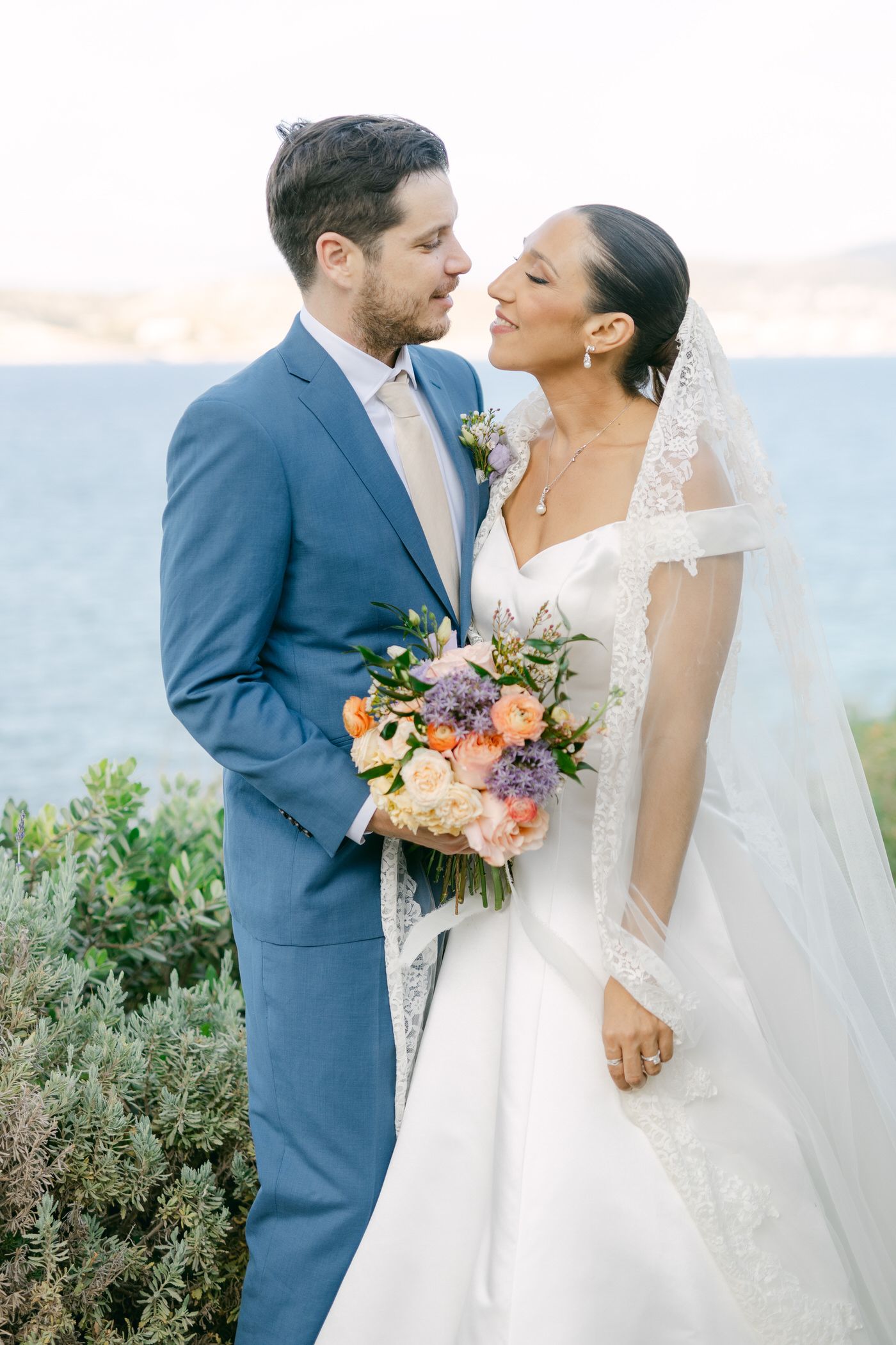 A wedding couple stands close together against an ocean backdrop, with the bride in white and groom in blue holding a bouquet.